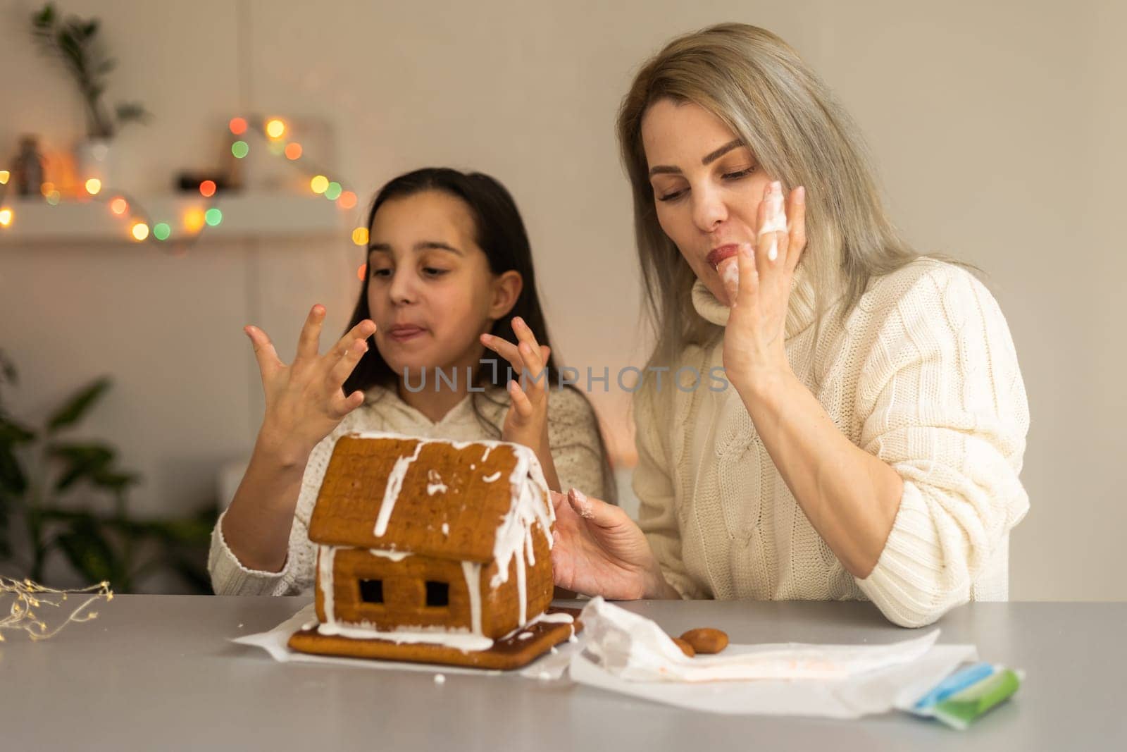 mother and daughter decorating gingerbread house. Beautiful living room with lights. Happy family celebrating holiday together. by Andelov13