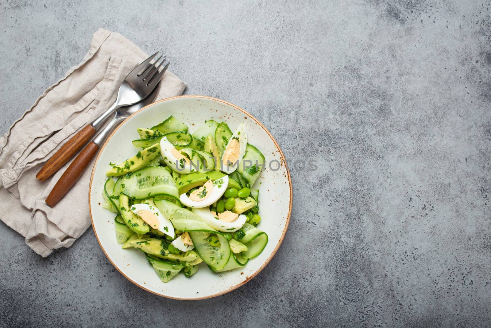 Healthy green avocado salad bowl with boiled eggs, sliced cucumbers, edamame beans, olive oil and herbs on ceramic plate top view on grey stone rustic table background. Space for text