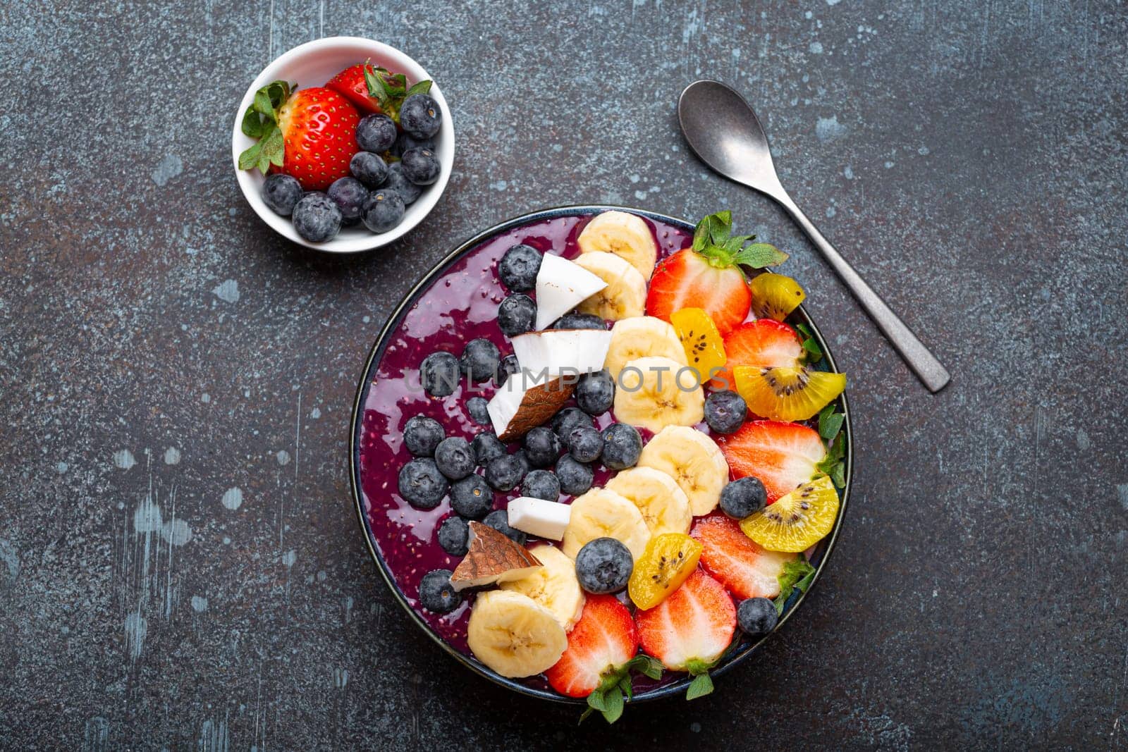 Healthy summer acai smoothie bowl with chia seeds, fresh banana, strawberry, blueberry, cocos, kiwi top view, rustic concrete background with spoon by its_al_dente