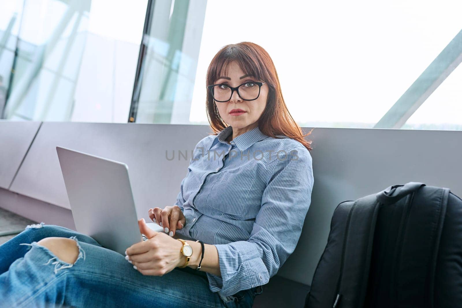 Middle aged woman with backpack waiting for airplane flight in airport terminal, using laptop, looking at camera. Travel, business trip, passenger air transport concept