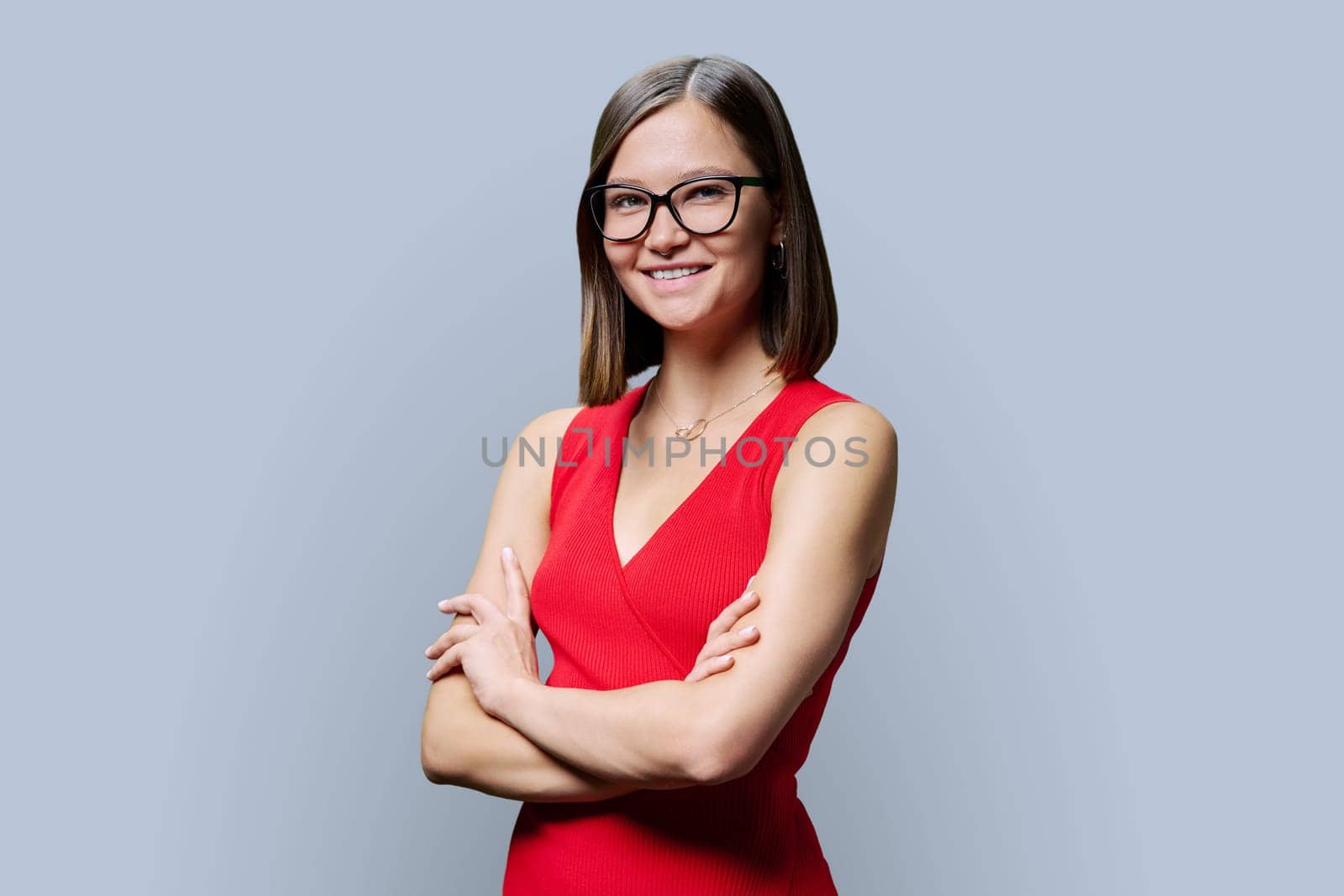 Young confident woman in glasses in red with crossed arms looking at camera on grey studio background. Business, work, services, education, fashion, beauty, professions, people