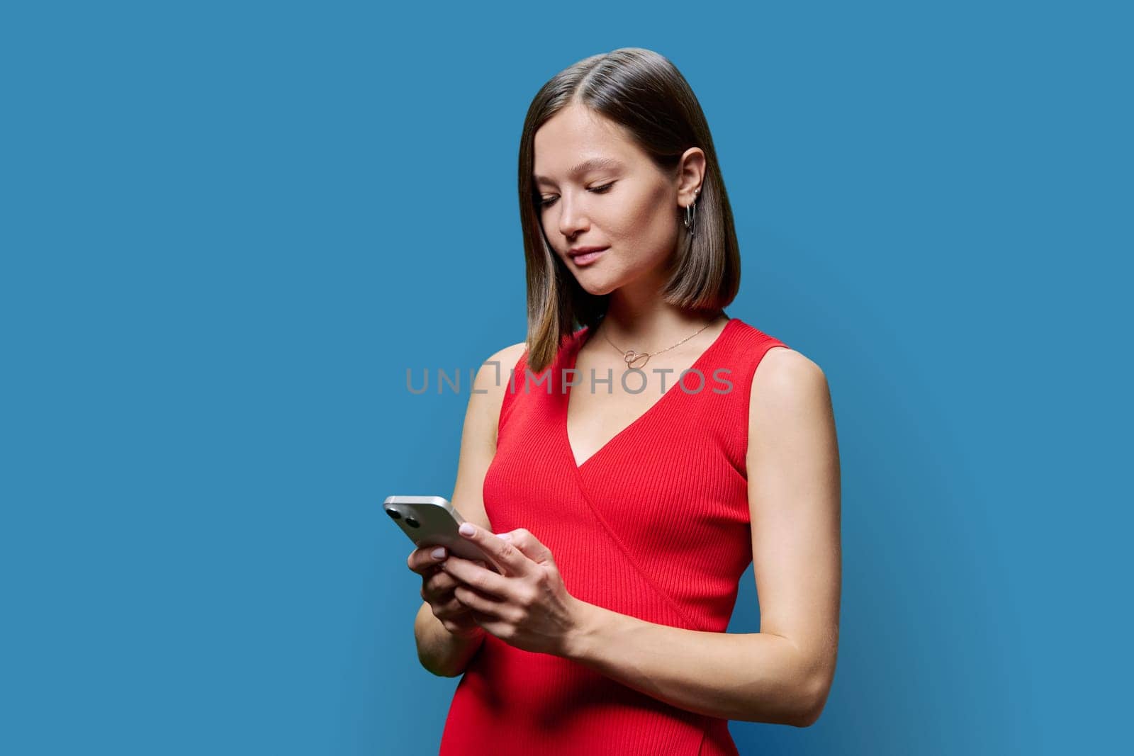 Young woman with smartphone in hands on blue studio background. Serious female looking at phone screen texting reading. Using mobile applications for work business shopping banking study leisure