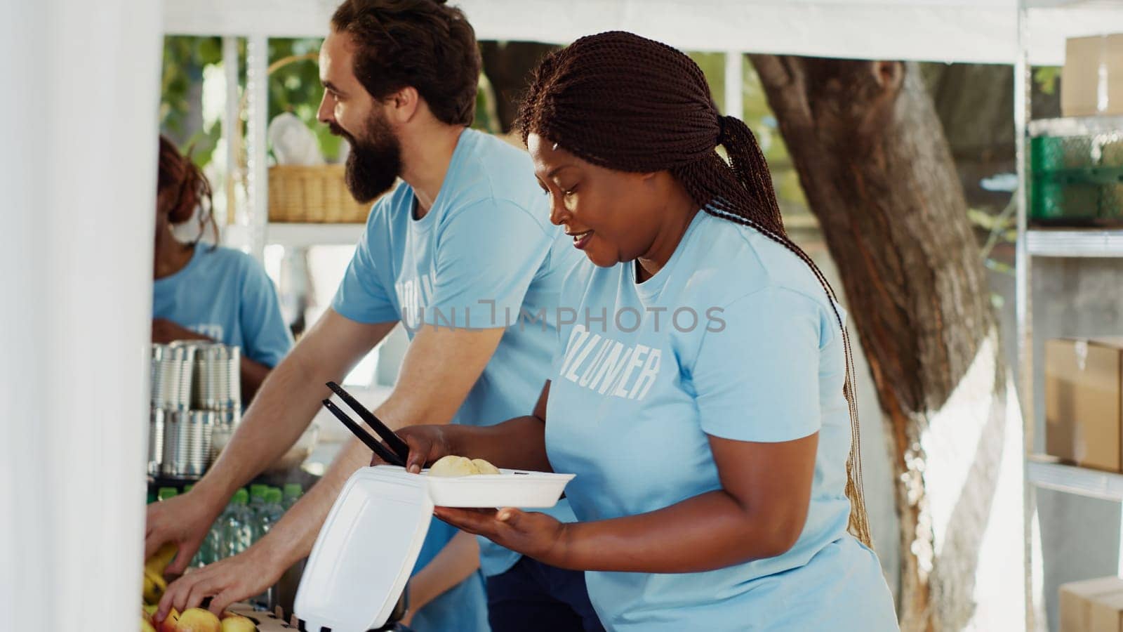 Black woman serving meals to underprivileged at community outreach event. Charity workers at a food drive distributing nutritious foods and essentials to the poor and homeless. Side-view, tripod shot.