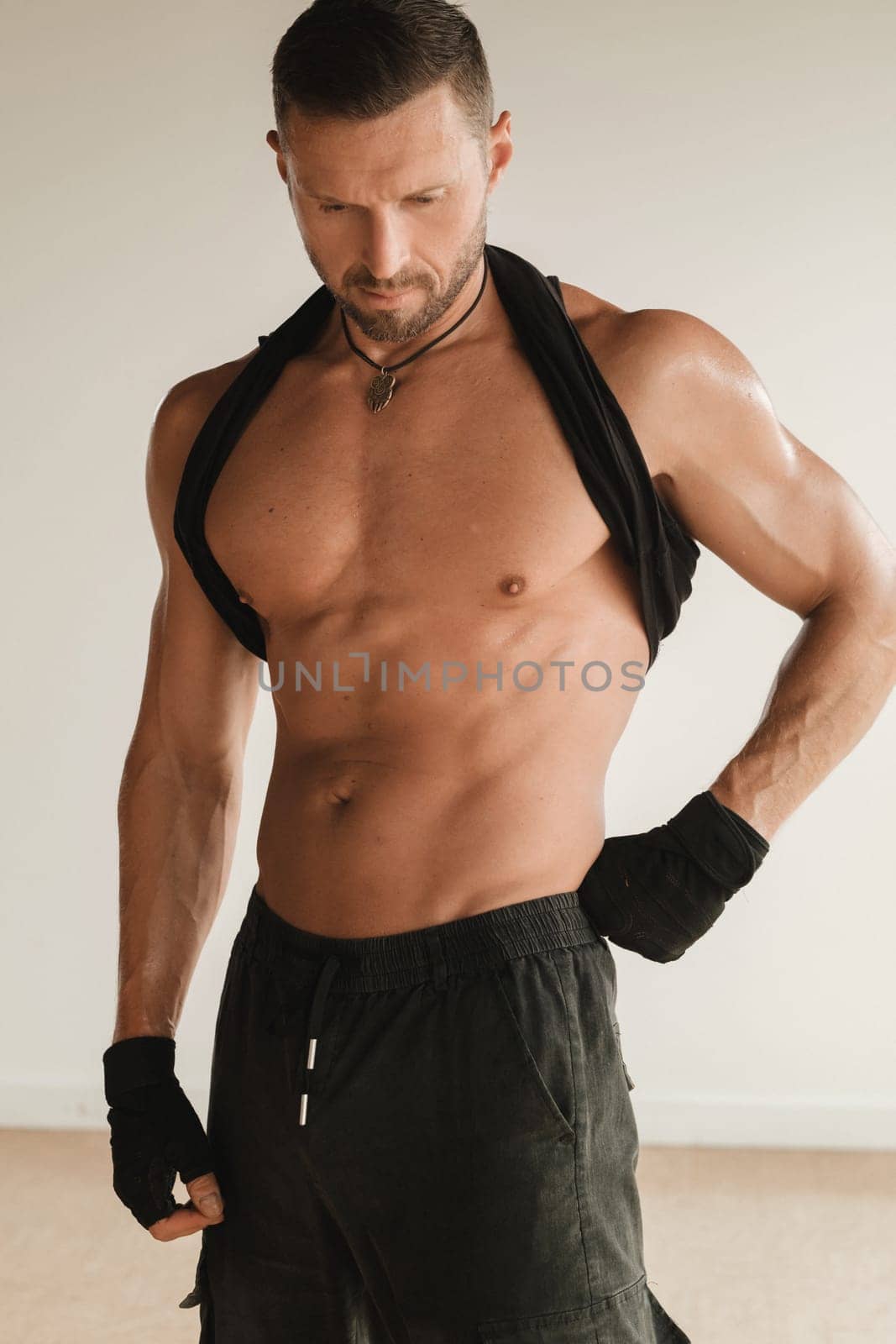 An adult muscular man with a naked torso stands in a room on a light background.