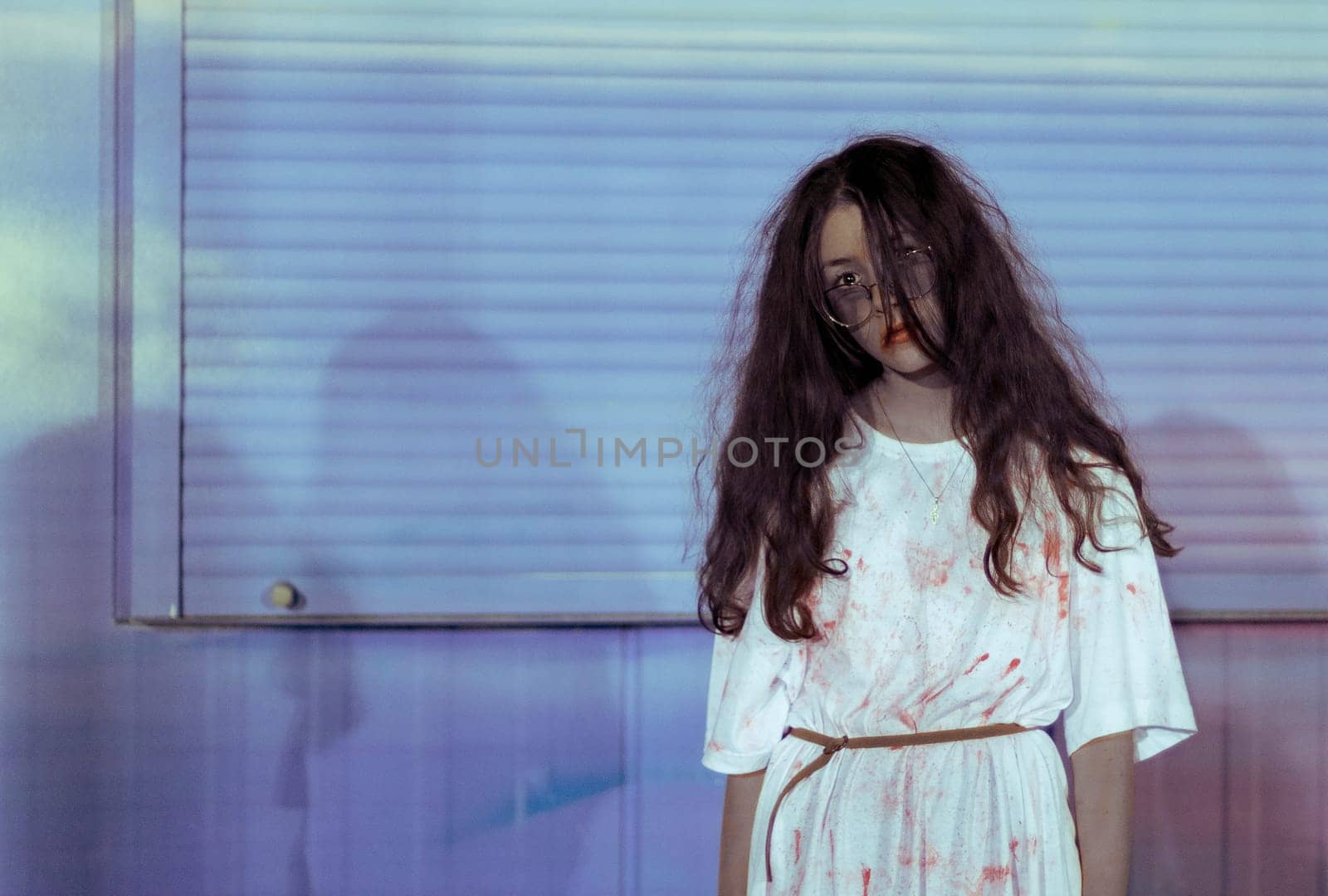 Portrait of a beautiful caucasian teenage girl in glasses, long flowing hair in a bloody white dress with a belt standing on the right against a background of a pale neon wall with copy space on the left, close-up side view.