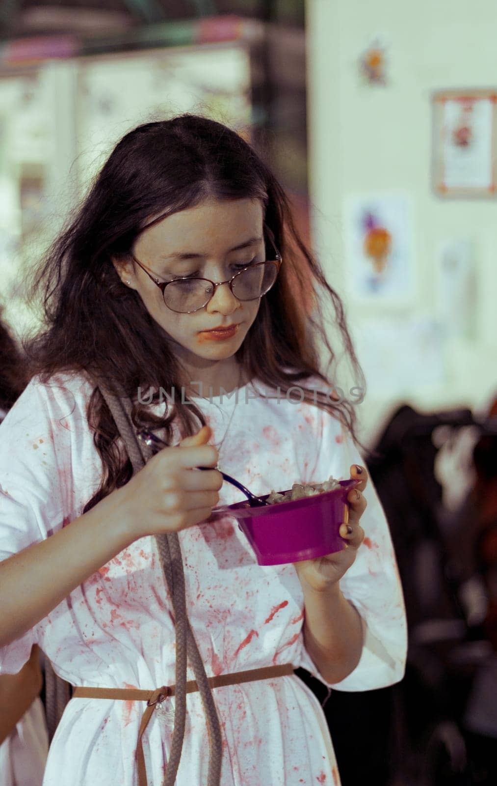 Portrait of a beautiful Caucasian teenager girl in a bloody white dress with ruffled long hair eats Halloween food from a bowl with a fork, standing on the street at night, close-up side view.