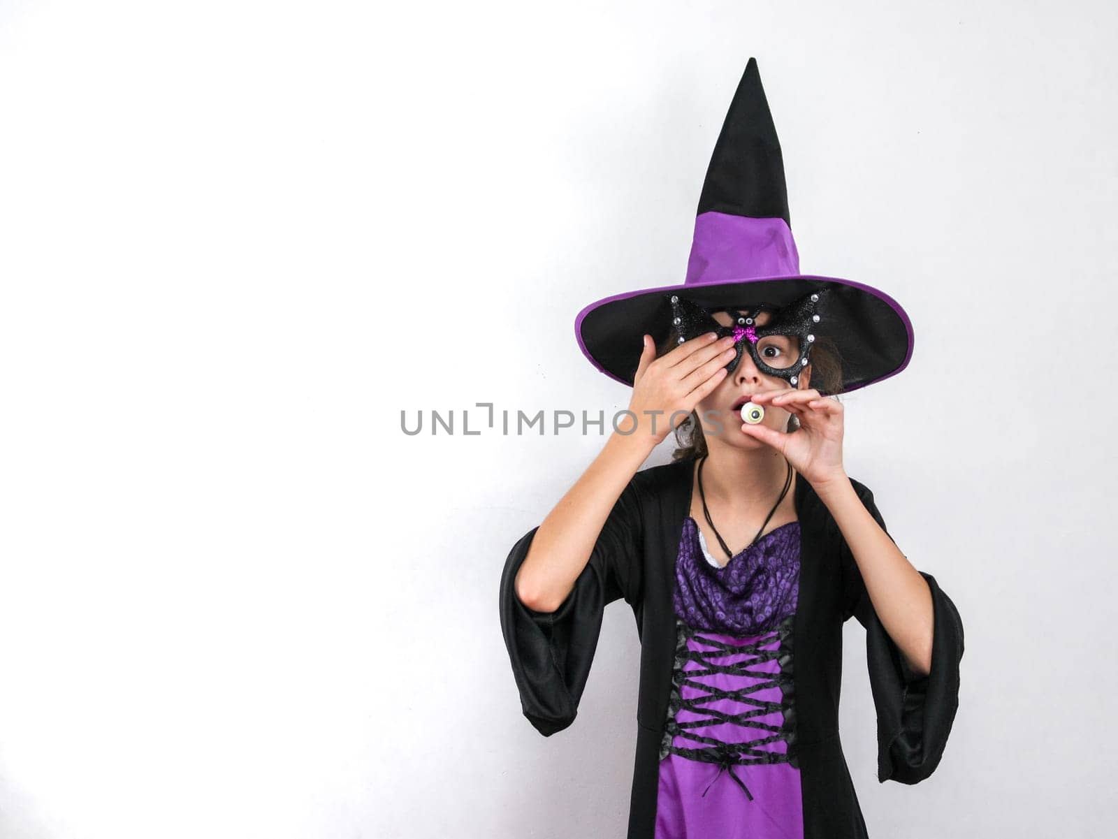 Portrait of a caucasian girl in a witch costume with a halloween hat holds candy near her mouth with one hand, and closes her eye with the other, stands on the right against a white wall with little copy space on the left, close-up side view. Halloween concept, Halloween celebration.