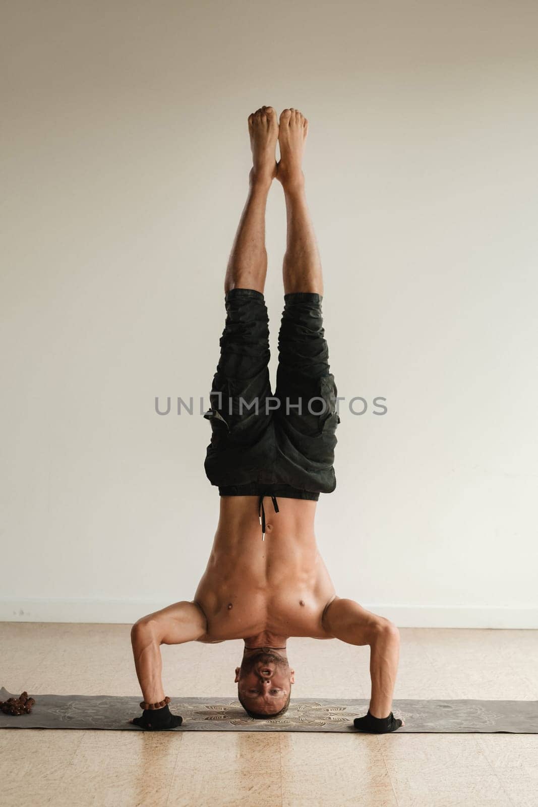 a man with a naked torso does yoga standing on his head indoors. Fitness Trainer by Lobachad