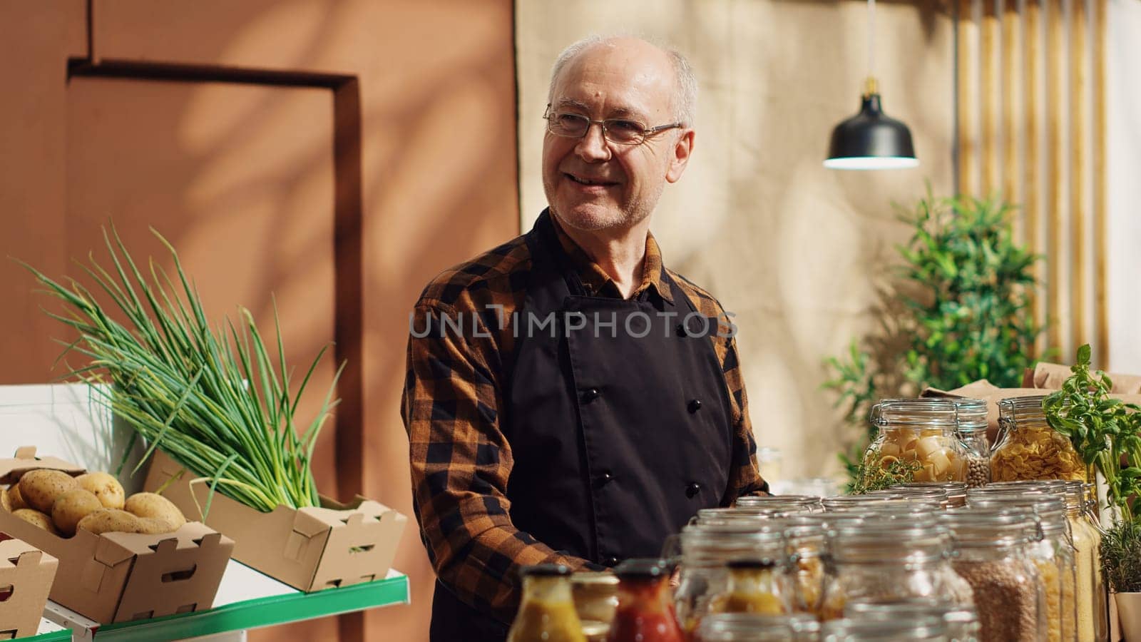 Portrait of smiling old zero waste supermarket owner using decomposable glass containers for its bulk items to lower climate impact. Elderly man in local grocery shop arranging merchandise