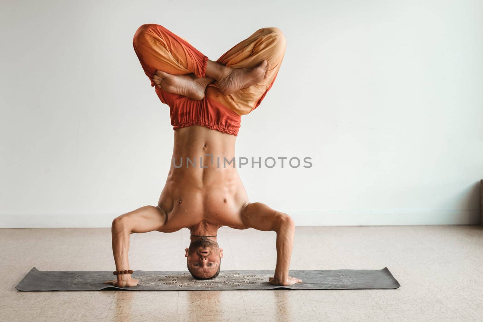 a man with a naked torso does yoga standing on his head indoors. Fitness Trainer by Lobachad