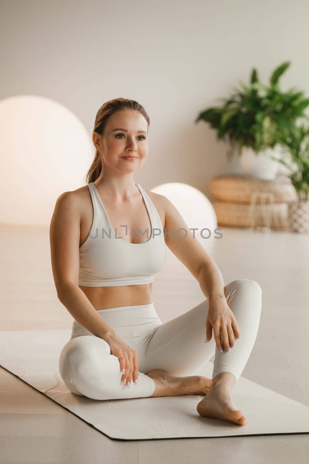 Portrait of a girl in white clothes sitting on a mat before doing Yoga indoors.