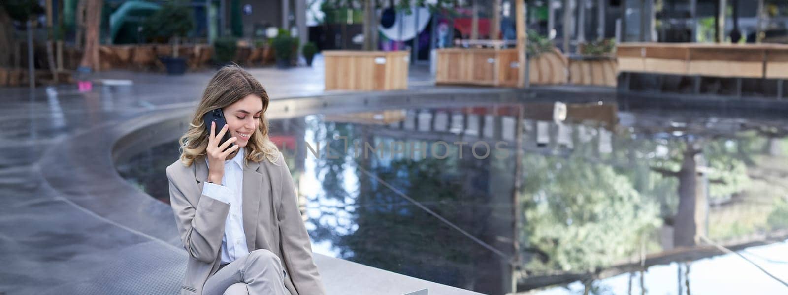 Vertical shot of corporate woman sitting outdoors with laptop, talking on mobile phone, working ouside office building while waiting for someone.