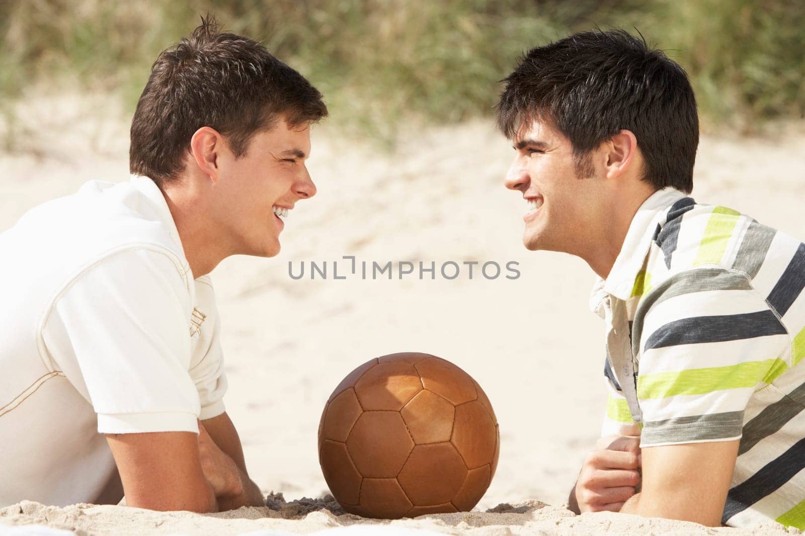 Two Young Men Relaxing On Beach With Football Together