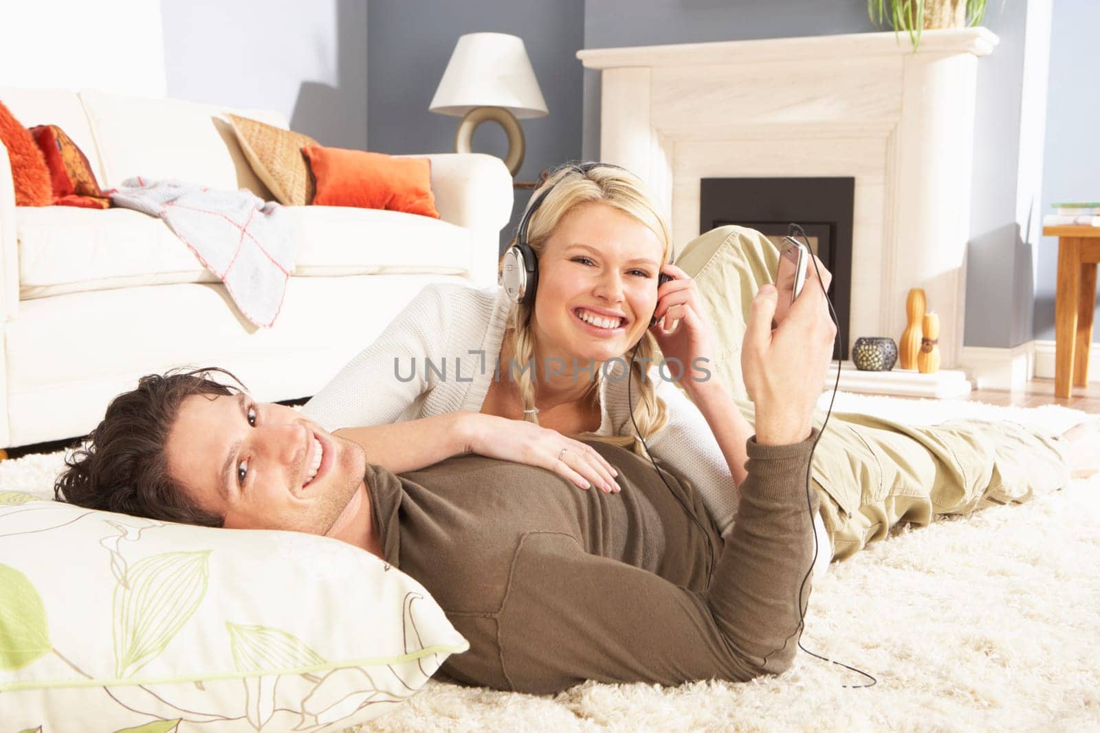 Couple Listening To MP3 Player On Headphones Relaxing Laying On Rug At Home