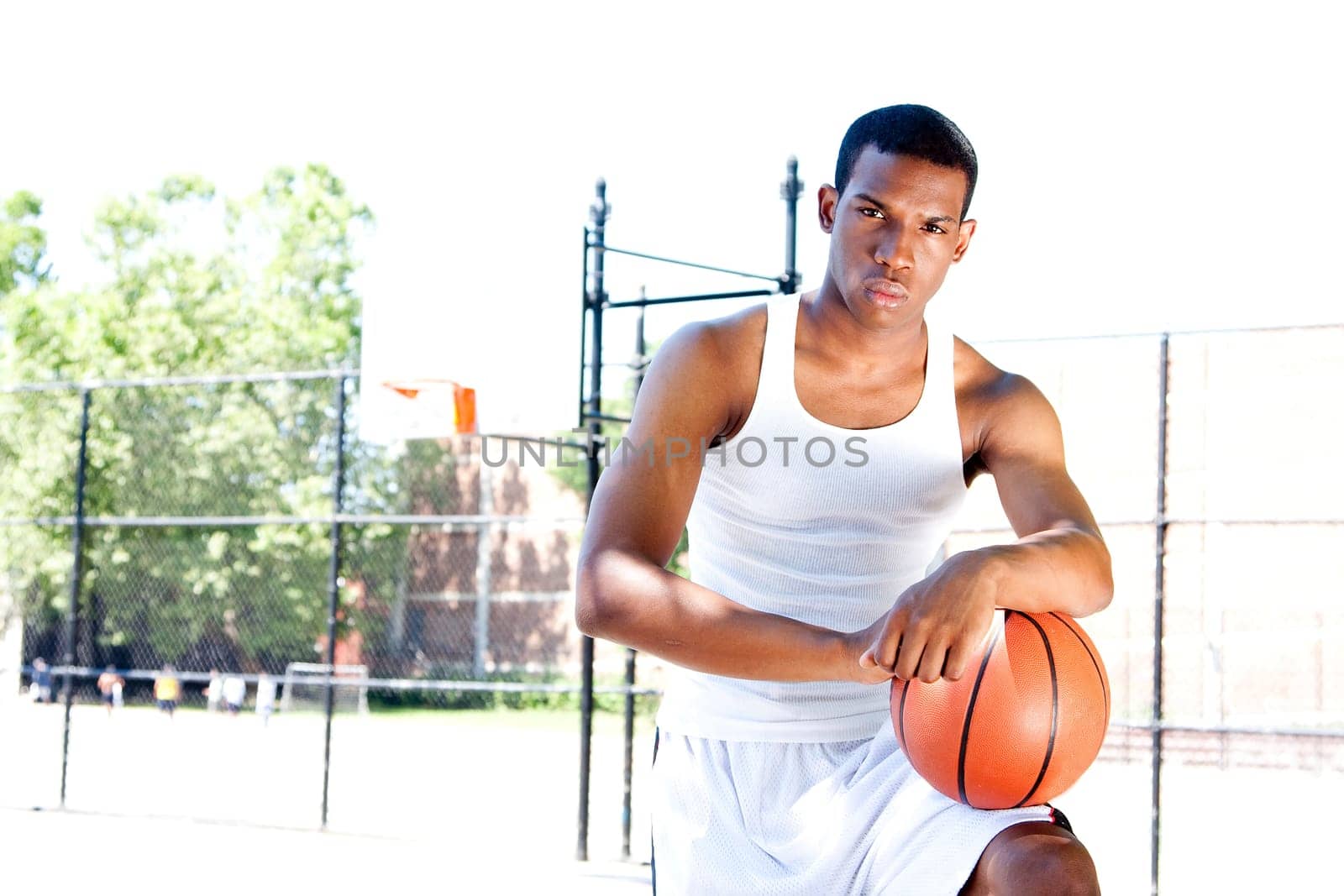 Handsome sporty African-American male basketball player with attitude dressed in white standing with his ball outdoor on a summer day in a basketball court.