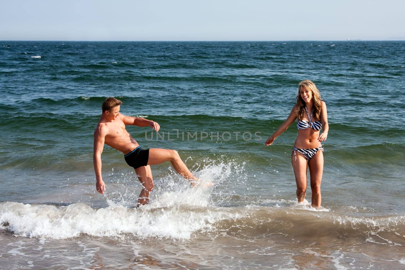 Beautiful young couple playing in the ocean splashing water, heaving fun on a summer day