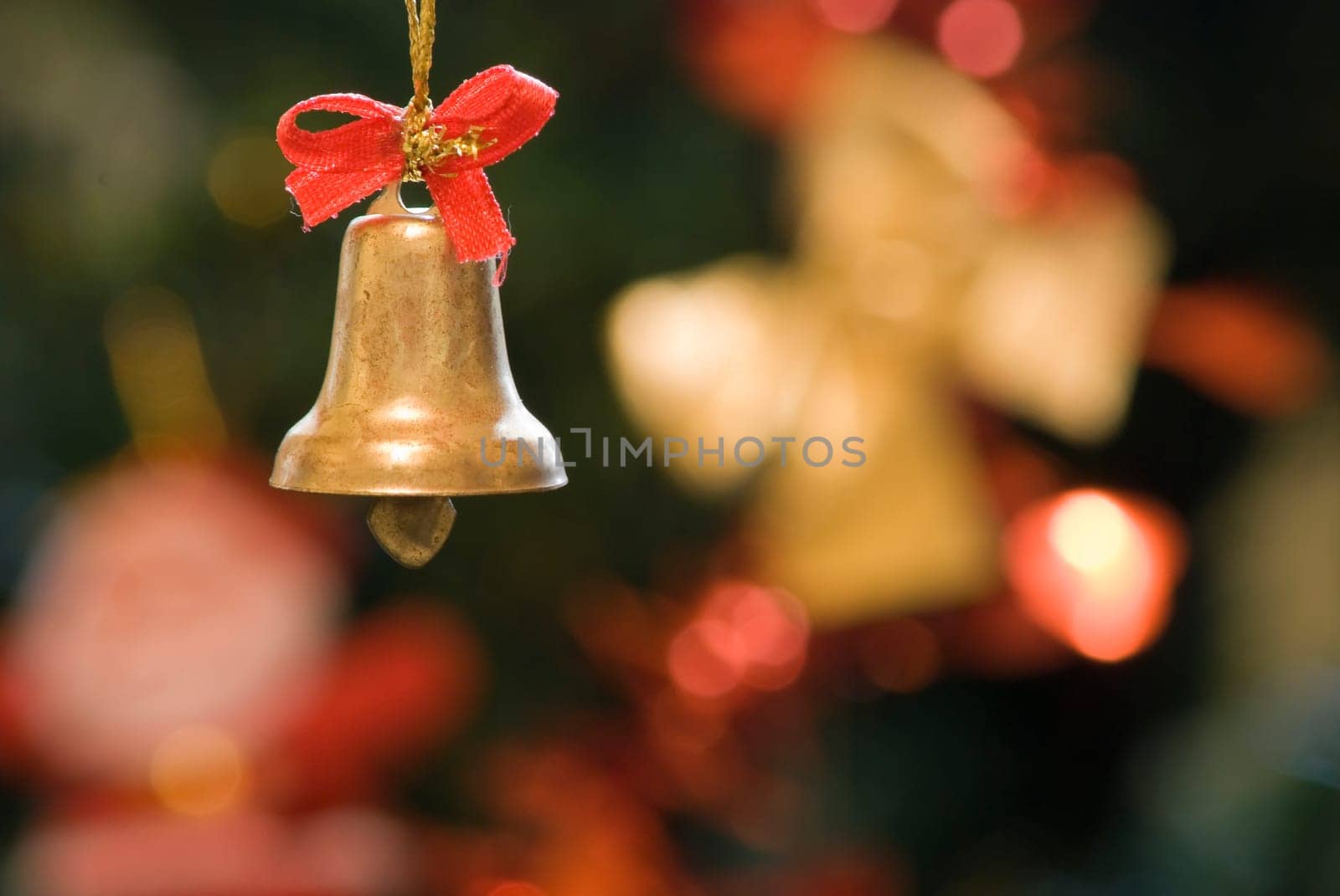 Decorative xmas bell with red ribbon with blurred xmas tree decoration in the background.