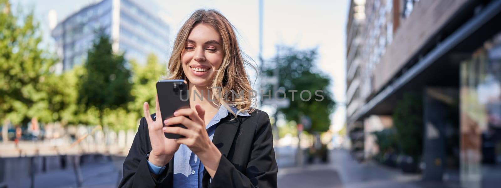 Portrait of smiling businesswoman using mobile phone while standing outdoors near office buildings by Benzoix