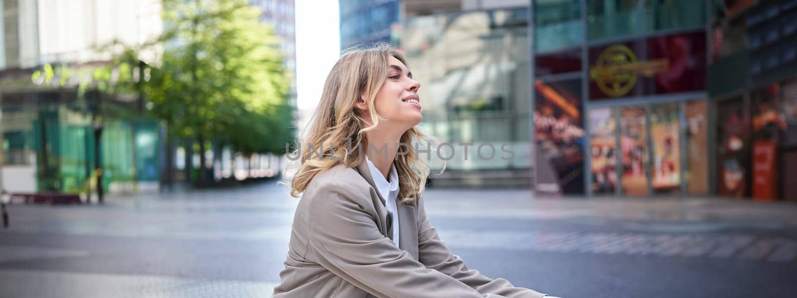 Smiling happy businesswoman in suit, sitting relaxed in city center and enjoying being outdoors by Benzoix