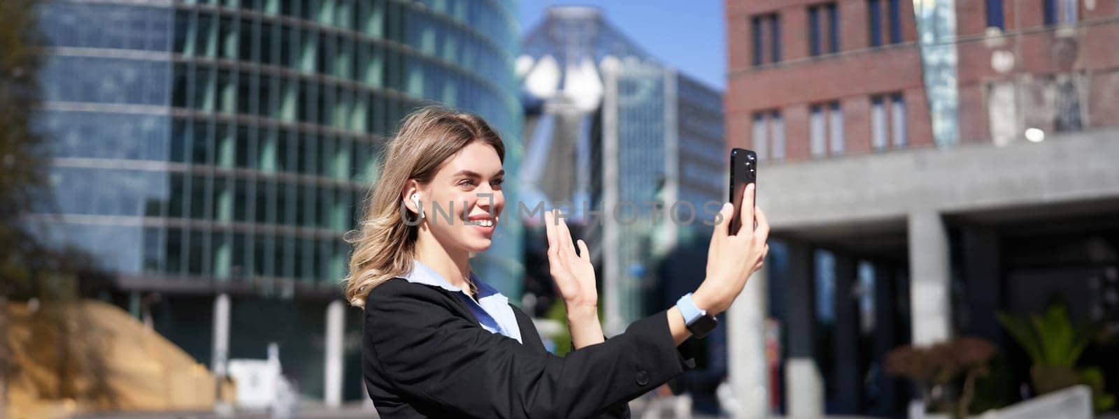 Portrait of businesswoman wave her hand at mobile phone camera, waves hand during video chat, stands in suit in city center outdoors by Benzoix