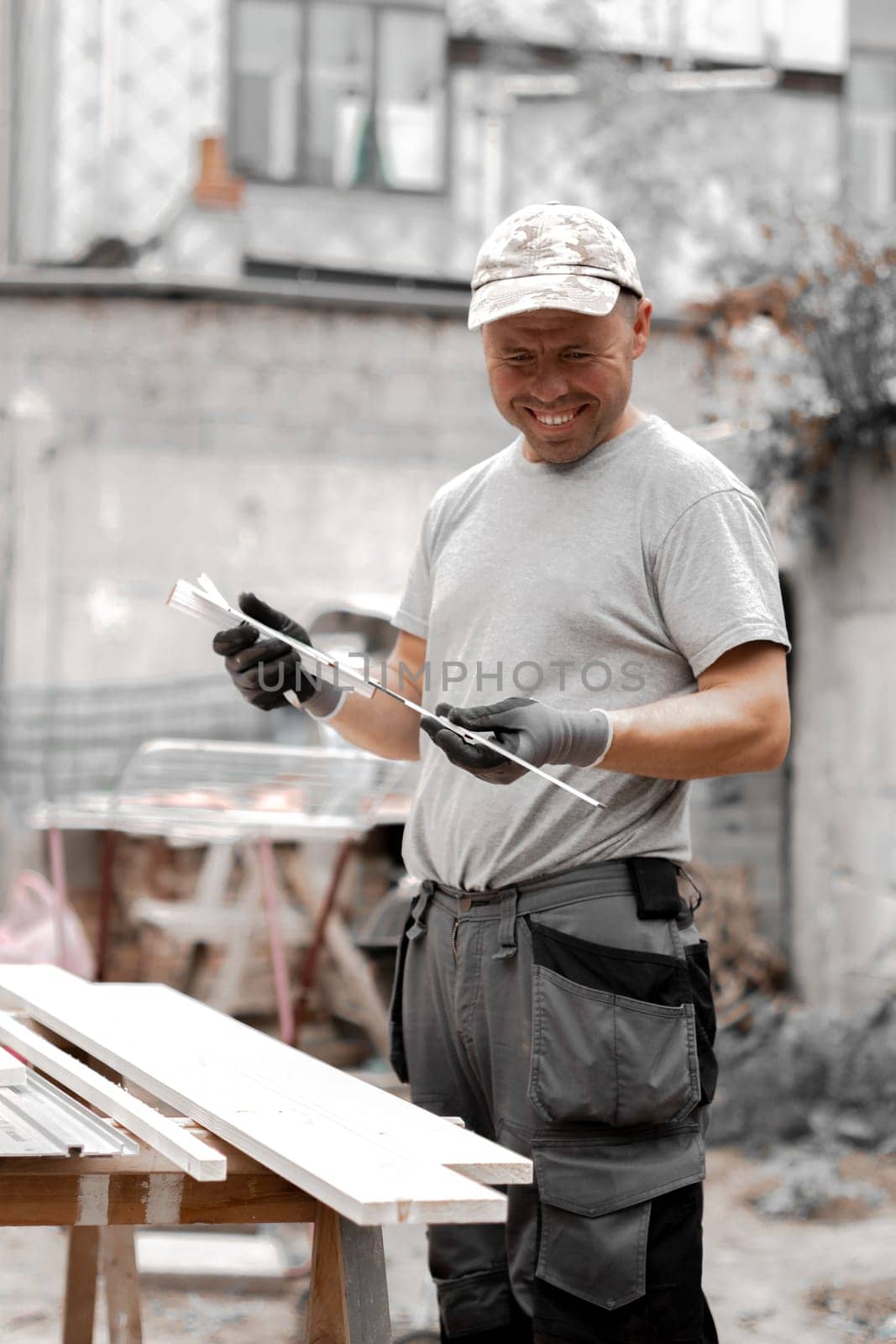 One young handsome Caucasian man in uniform and gray gloves with a happy smile holds a construction measuring tape and prepares to draw on a board with a pencil, standing in the backyard of a house on a summer day, close-up side view.