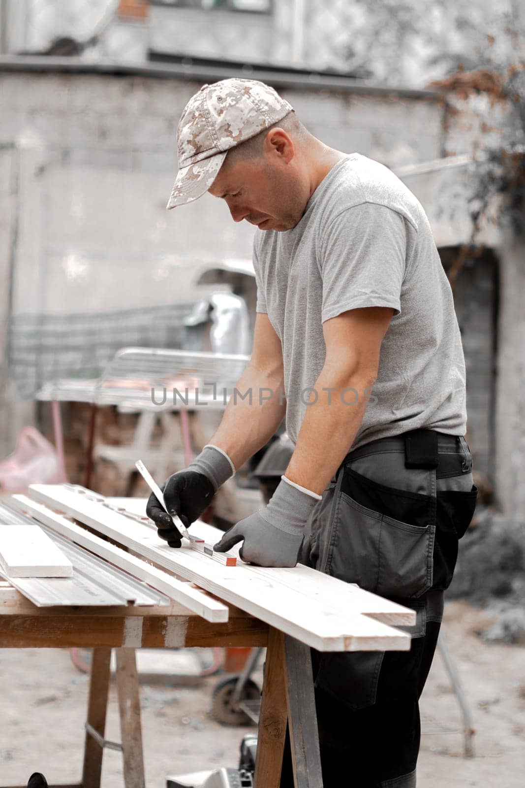 One young handsome Caucasian man in a uniform and gray gloves stands thoughtfully half sideways and draws with a pencil, preparing to cut a board in the backyard of a house on a summer day, close-up side view.