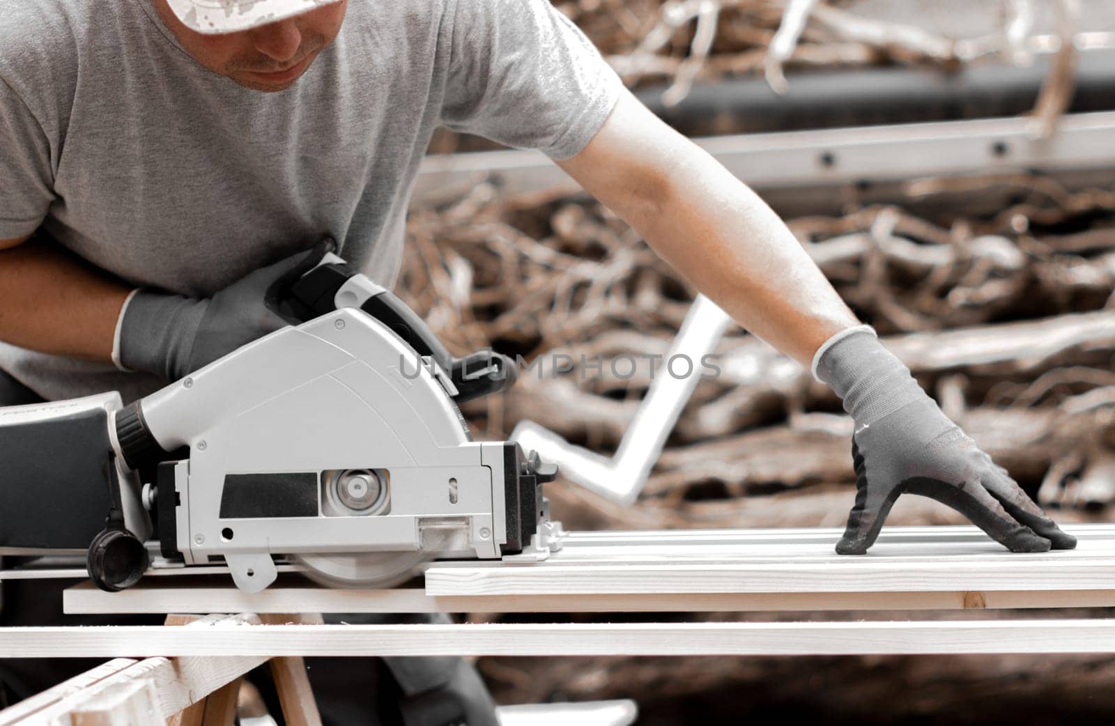 One young Caucasian recognizable man in a uniform and gray gloves begins to saw a board into beams with an electric saw, standing in the backyard of a house on a summer day, close-up side view.