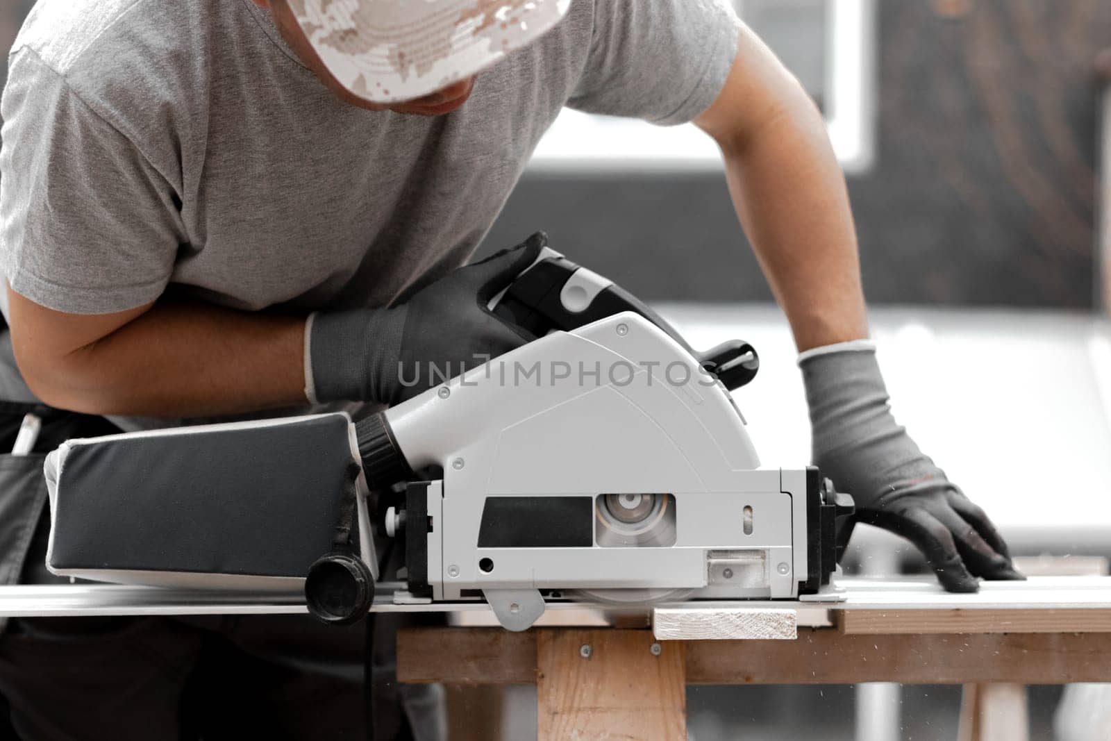 One young Caucasian recognizable man in a uniform and gray gloves is sawing a board into beams with an electric saw, standing in the backyard of a house on a summer day, close-up side view.