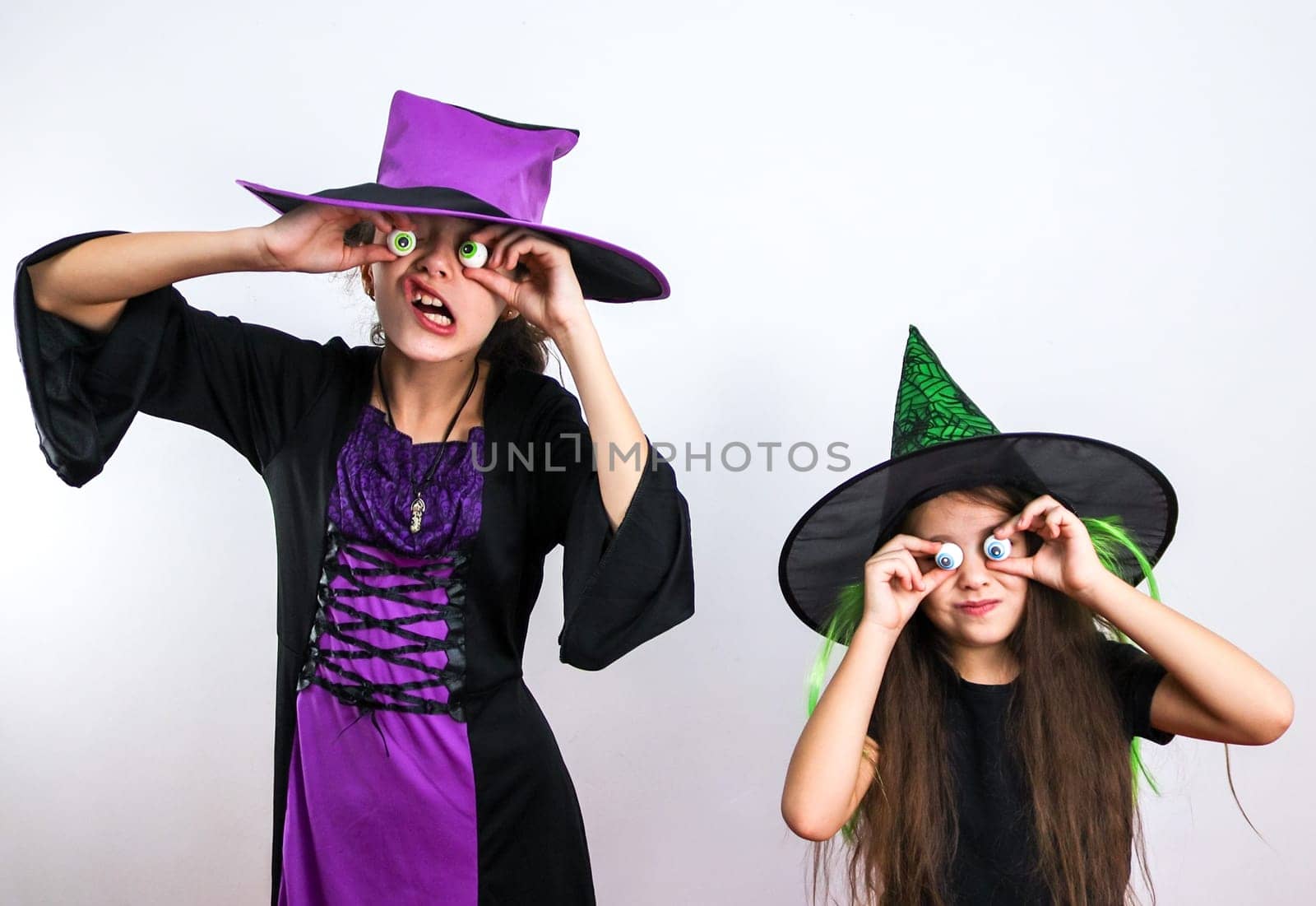 Two caucasian girls in witch costumes hold candies on their hands in front of their eyes and express halloween emotions, close-up side view.