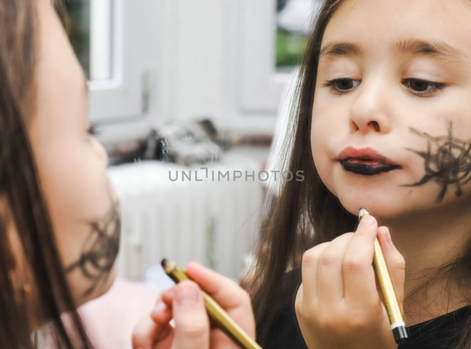 Caucasian brunette girl with brown eyes and black painted cobwebs on her cheek paints her lips with a pencil, looking in the mirror and getting ready for the celebration of Halloween, close-up side view. Halloween preparation concept, halloween makeup.