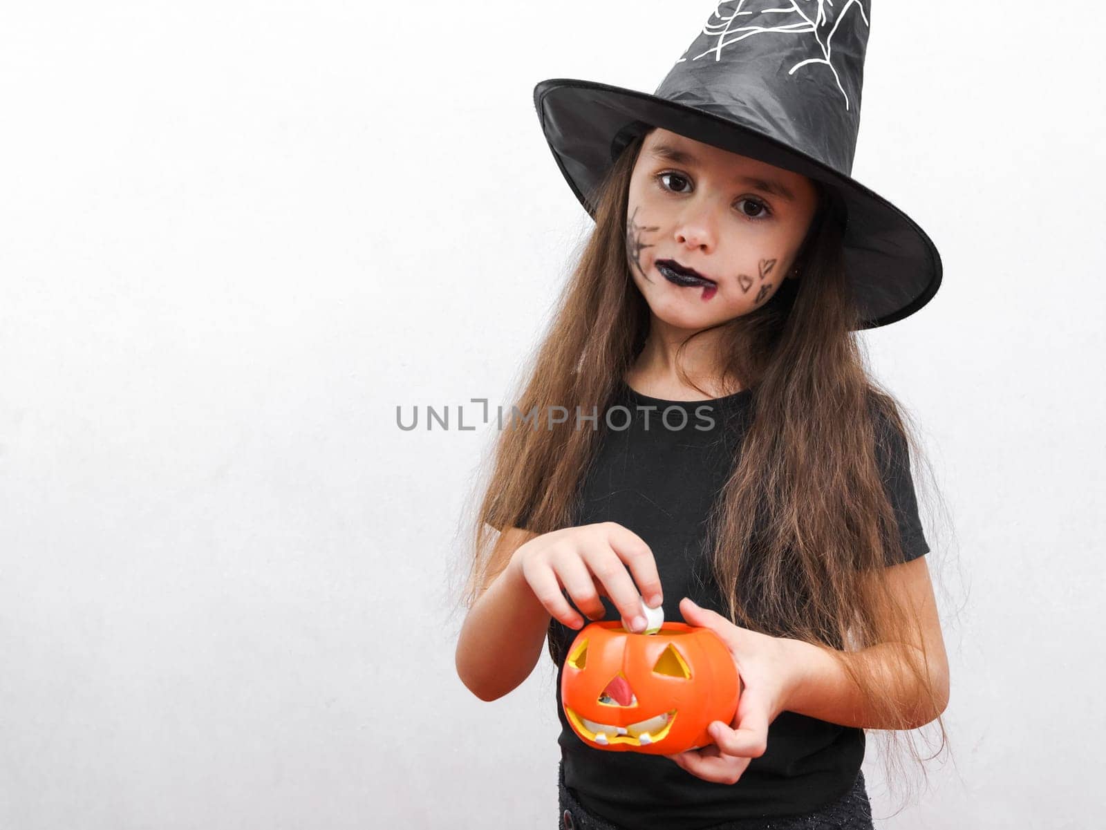 Portrait of a beautiful caucasian brunette girl in black clothes with long hair, halloween makeup and a witch hat holds a ceramic pumpkin with candies in her hands looking at the camera. stands on the right on a white background with copy space on the left, close-up side view. Halloween concept, Halloween celebration.