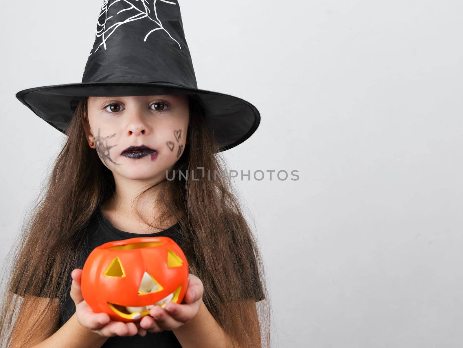 Portrait of a beautiful caucasian brunette woman in black clothes with long hair, Halloween makeup and a witch hat holds a ceramic pumpkin with candies in her hands, looking at the camera, standing on the left on a white background with a small copy space on the right, close-up side view. Halloween concept, Halloween celebration.