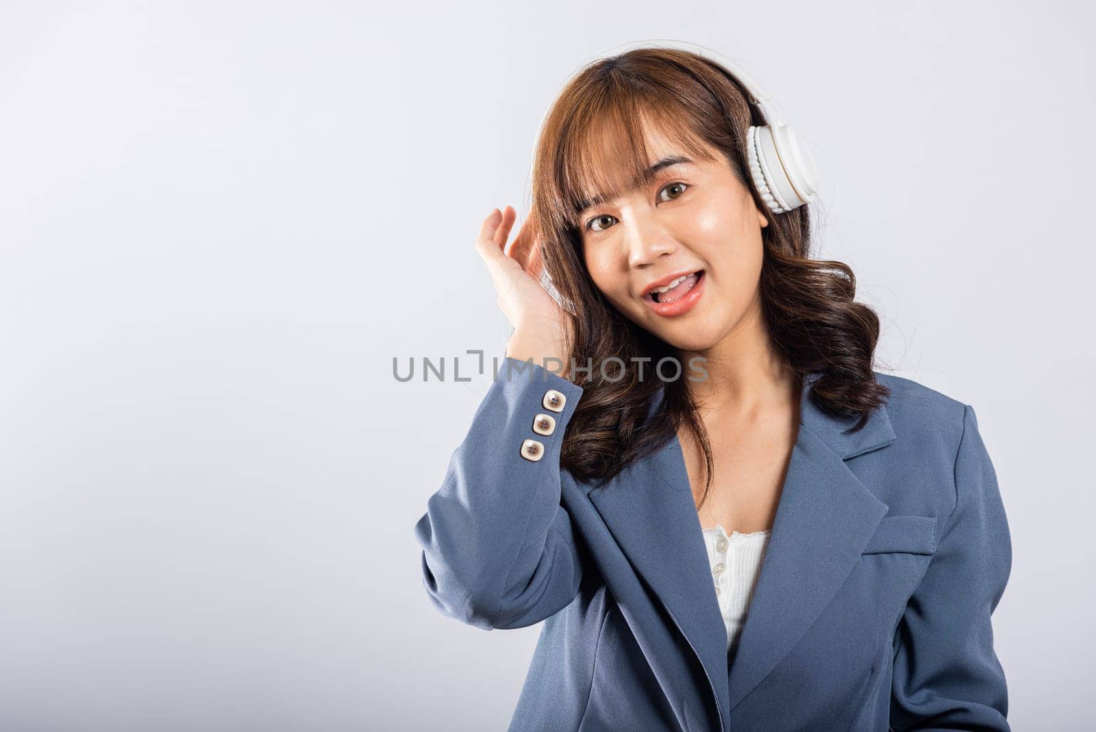 Excited woman in Bluetooth headphones smiles, immersed in music from her phone's radio. Studio shot, isolated on white. This happy Asian young female embraces modern technology for her enjoyment.