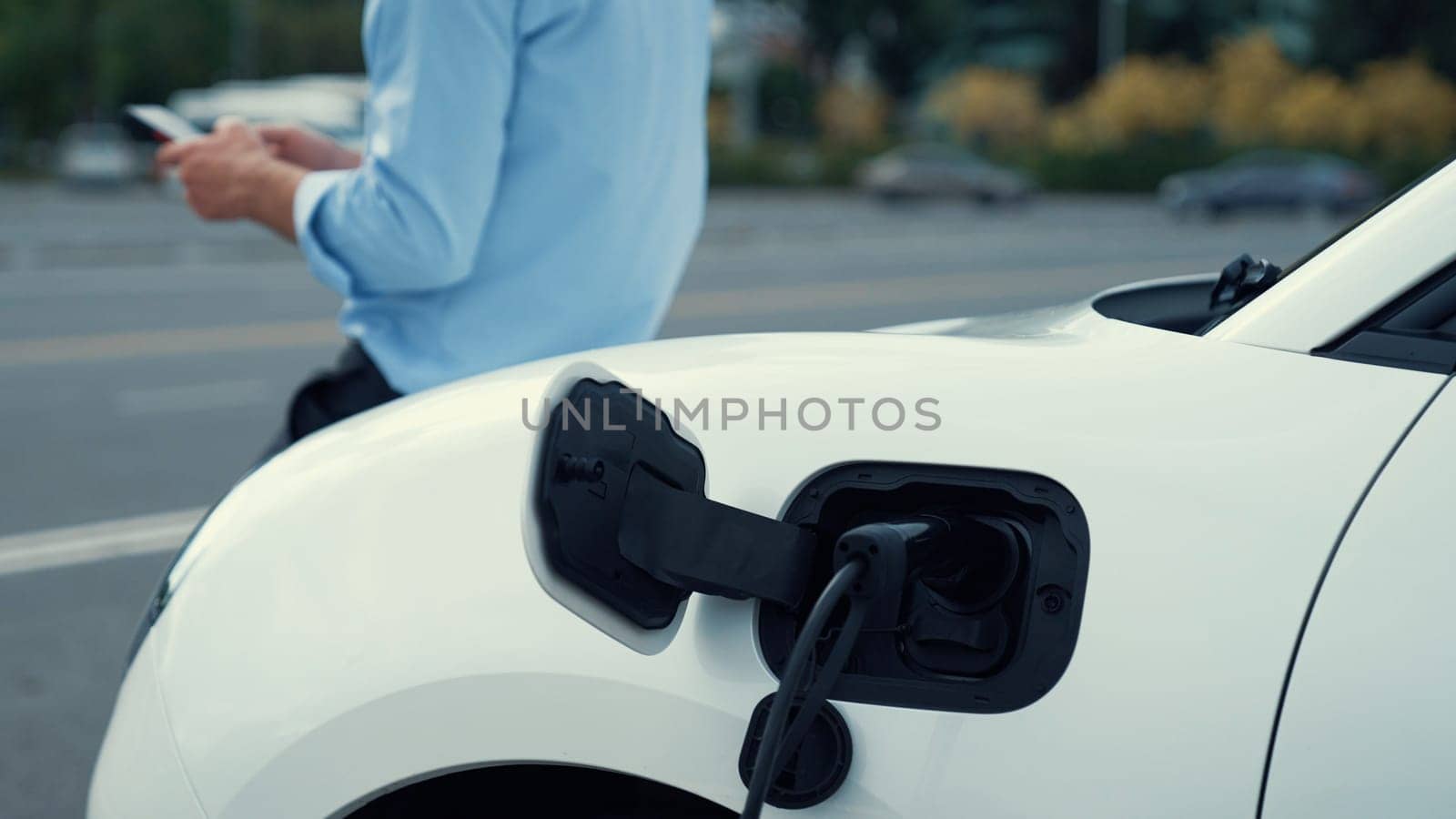 Suit-clad businessman with progressive ambition leaning on his electric vehicle while standing on a charging station with a power cable plug and a renewable energy-powered electric vehicle.