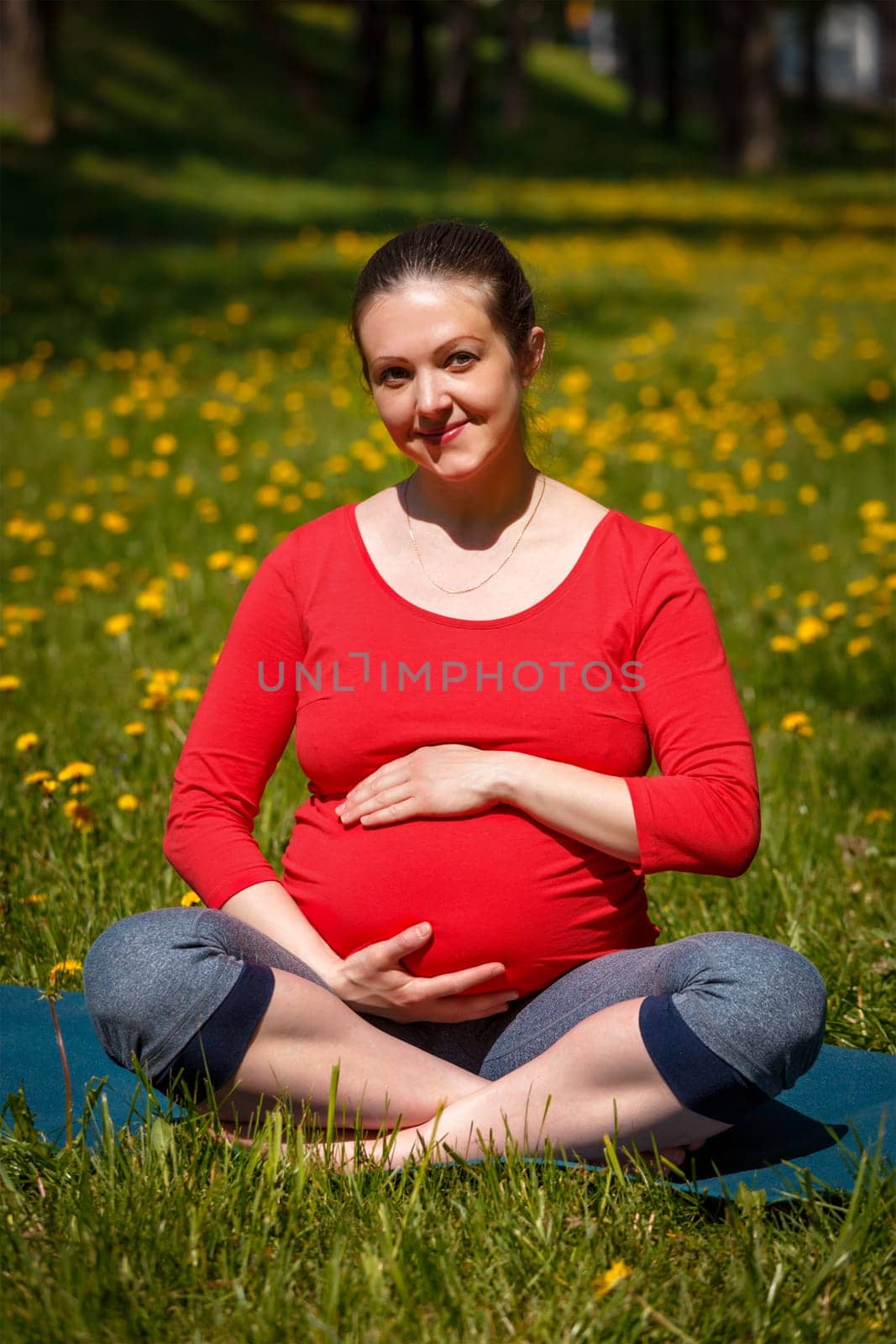 Pregnancy yoga exercise - pregnant smiling woman doing asana Sukhasana easy yoga pose holding her abdomen outdoors on grass lawn with dandelions in summer
