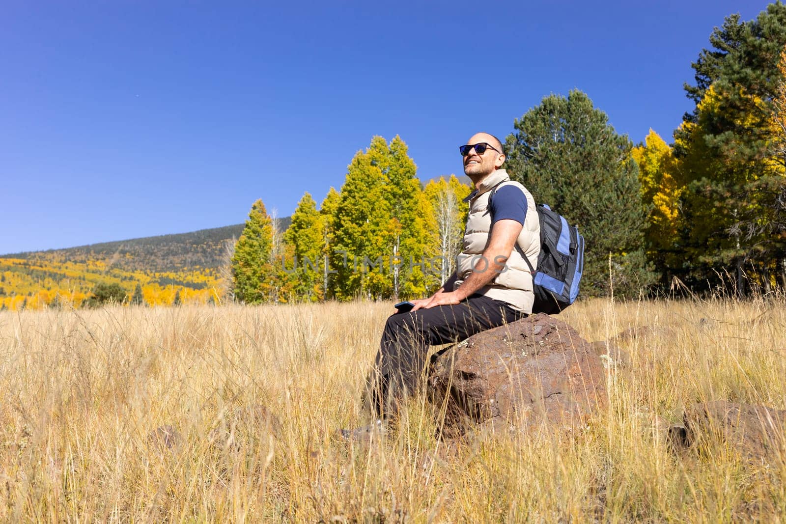 Happy Smiling Man Takes A Rest During Hiking In Mountains At Fall Season On Sunny Day. Male Hiker with Backpack Does Sport , Trekking In Autumn Nature On Vacation. Travel, Active Lifestyle. Copy Space