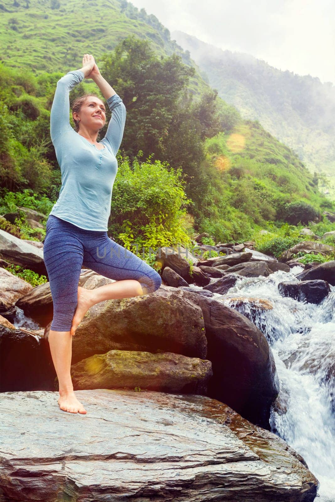 Woman in yoga asana Vrikshasana tree pose at waterfall outdoors by dimol