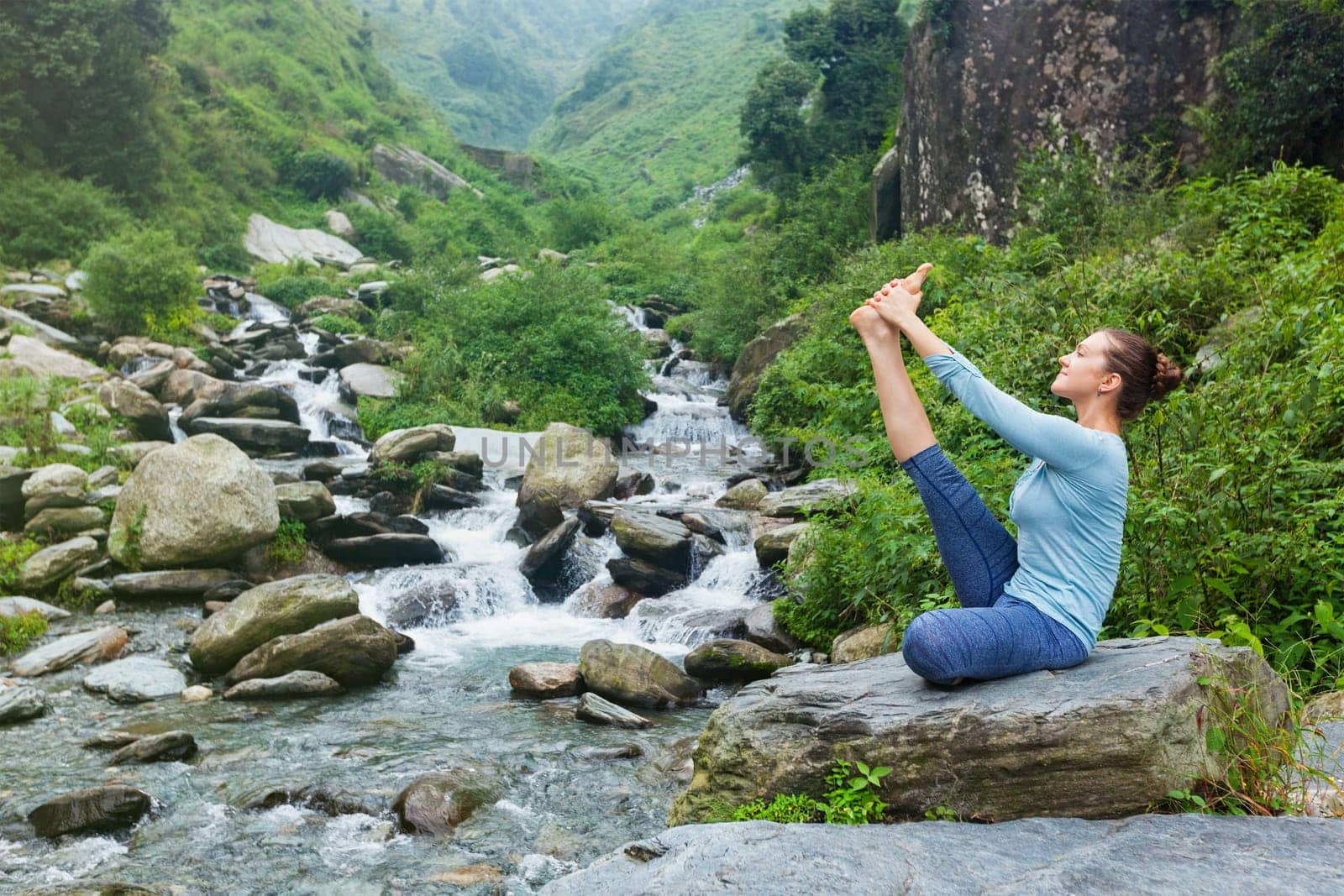 Yoga exercise outdoors - woman doing Ashtanga Vinyasa Yoga balance asana Ubhaya padangusthasana Big Double Toe Yoga Pose at tropical waterfall in Himalayas in India