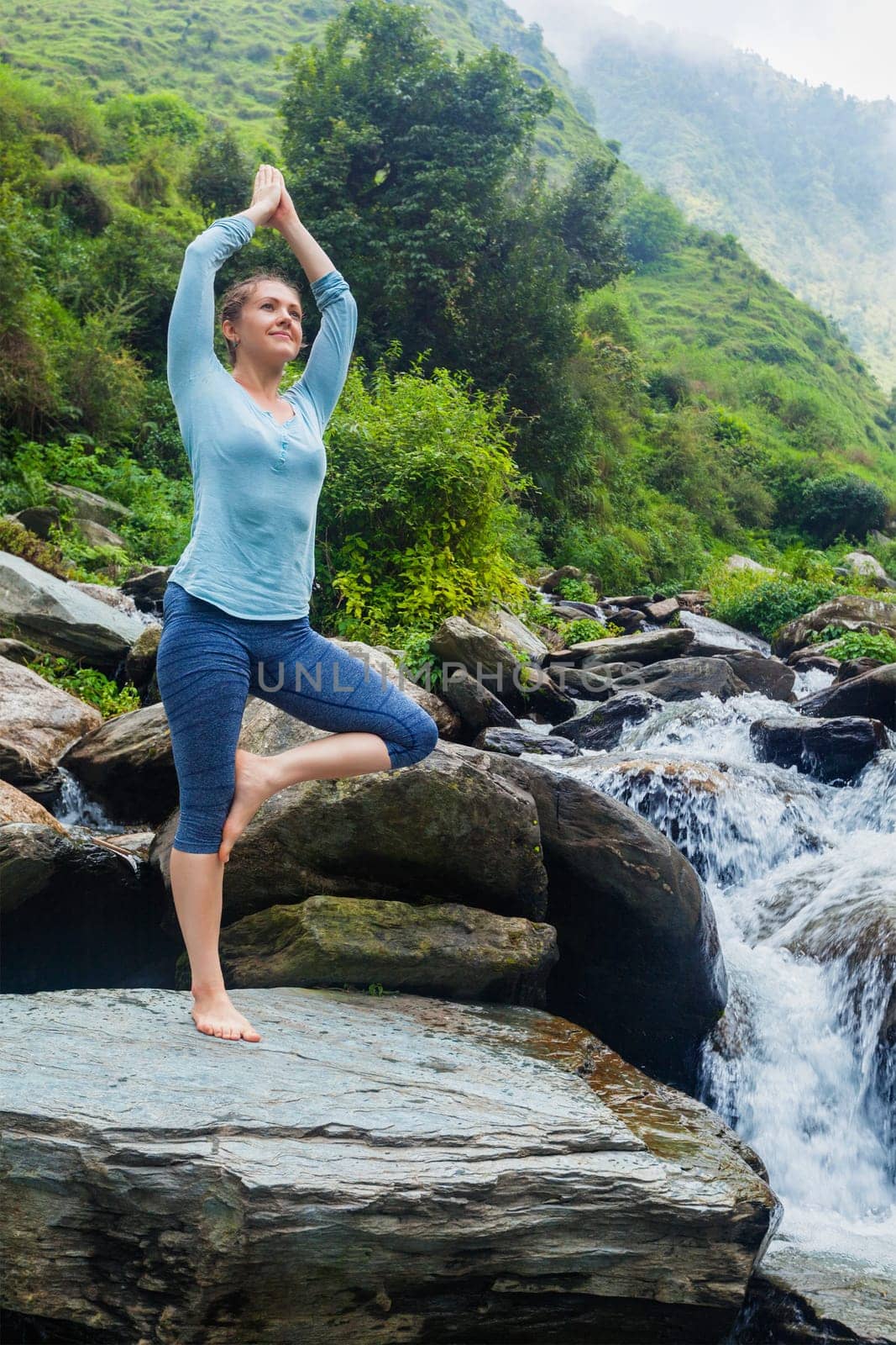 Woman in yoga asana Vrikshasana tree pose at waterfall outdoors by dimol