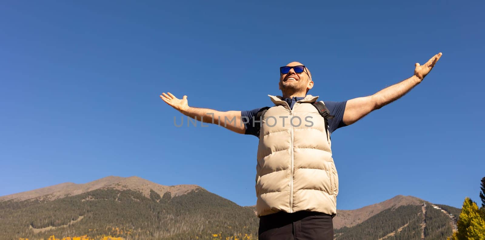 Banner Handsome 40 yo Male Tourist with Open Arms, Mountains, Blue Sky on Background. Male Hiker with Backpack Does Hiking, Trekking In Autumn Nature. Travel, Active Lifestyle Copy Space. Horizontal by netatsi