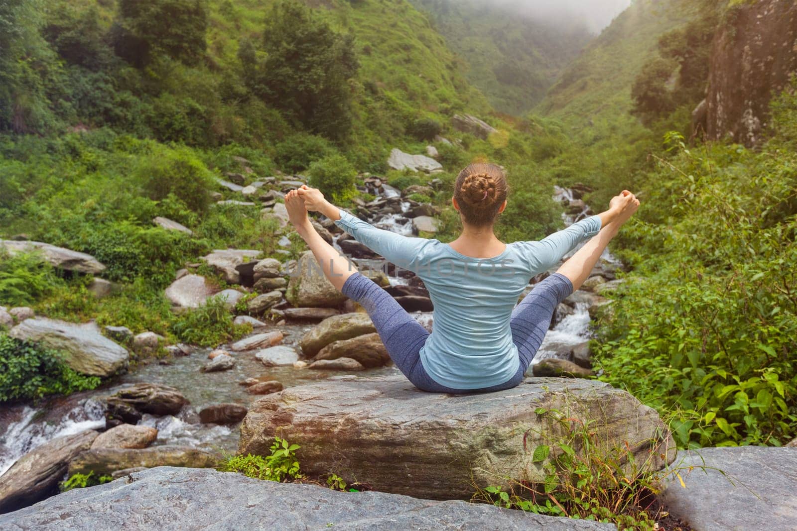 Woman doing Ashtanga Vinyasa asana Upavistha Konasana outdoors at tropical waterfall