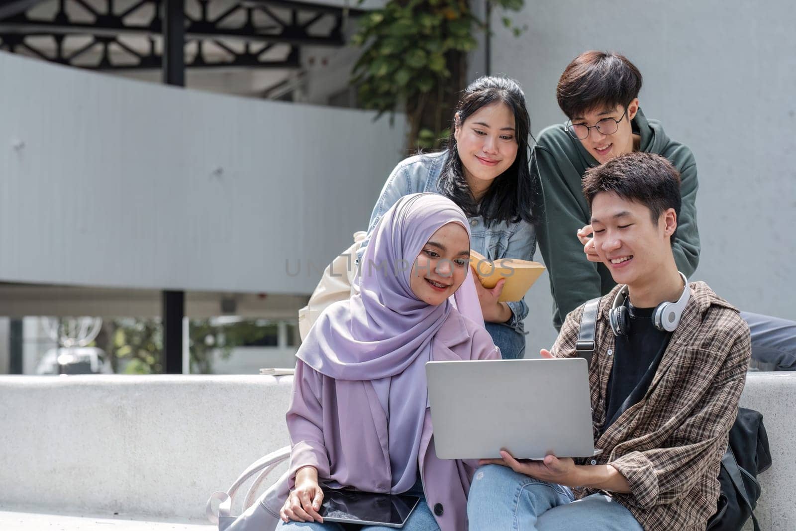 Group of happy young Asian college students sitting on a bench looking at a laptop screen, discussing and brainstorming on their school project together by wichayada