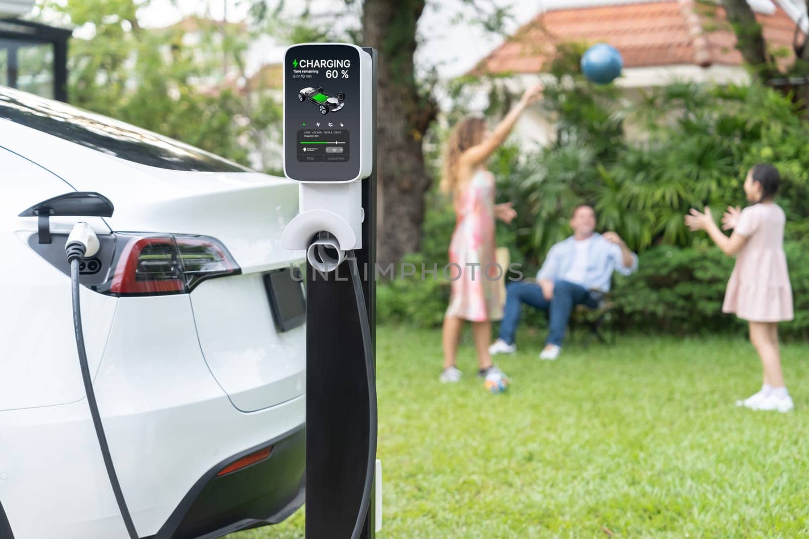 Focus electric vehicle recharge from home charging station on blur background of happy and playful family playing together. EV car using alternative and sustainable energy for better future.Synchronos