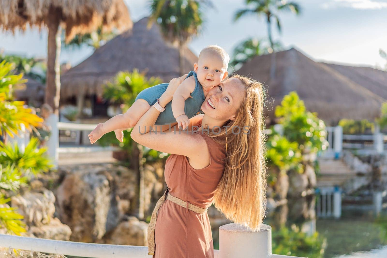 Amidst tropical palms and thatched roofs, a loving mom embraces her baby, sharing warmth and affection in a tranquil outdoor setting by galitskaya