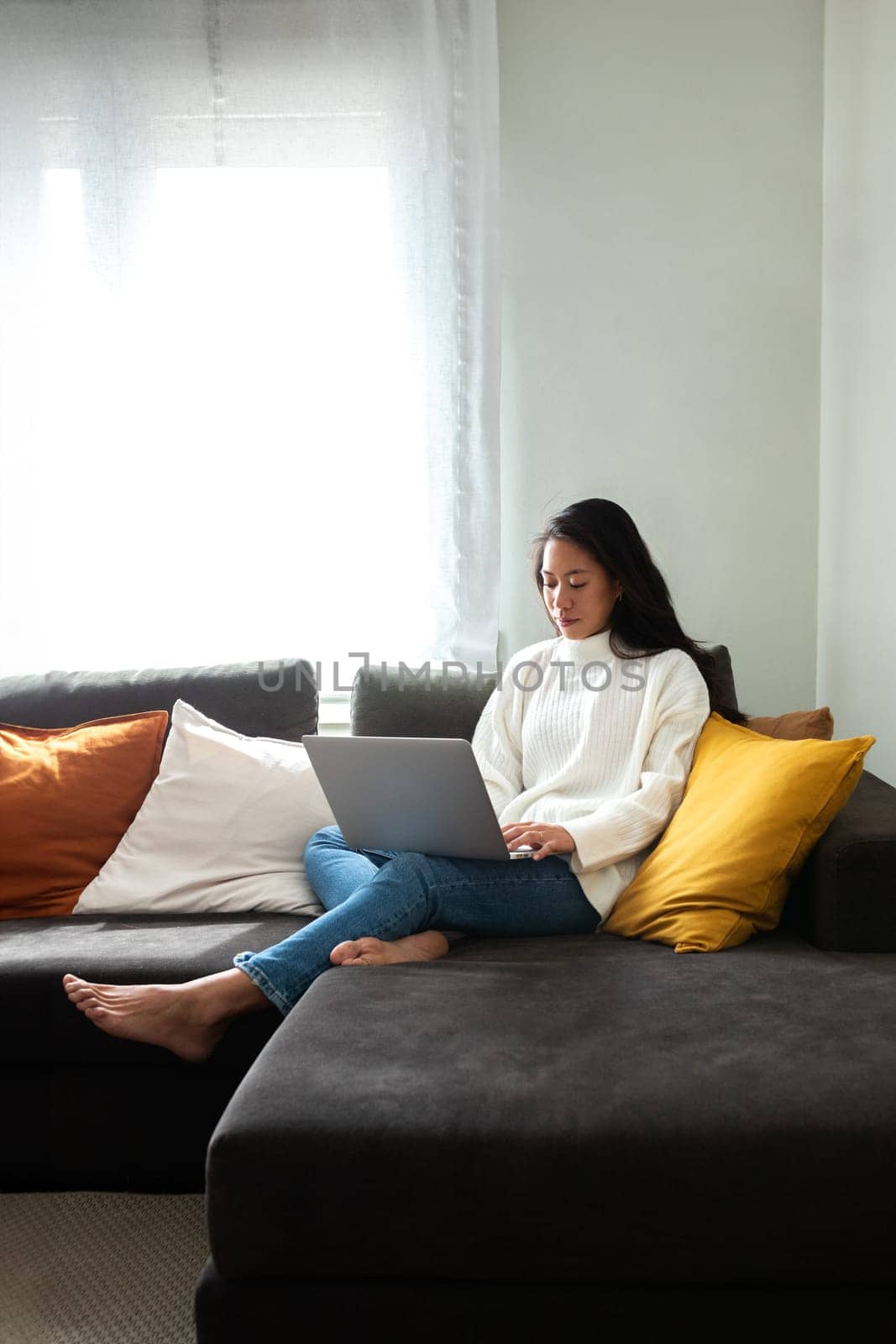 Young Asian woman working with laptop at home sitting on the couch in cozy apartment living room. Copy space. by Hoverstock