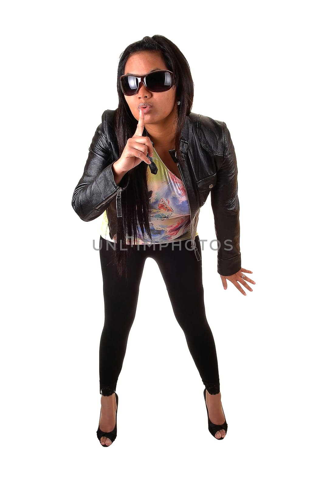 An Asian woman in a black leather jacket and tights, with sunglasses holds her finger for her mouth, be quiet, for white background.