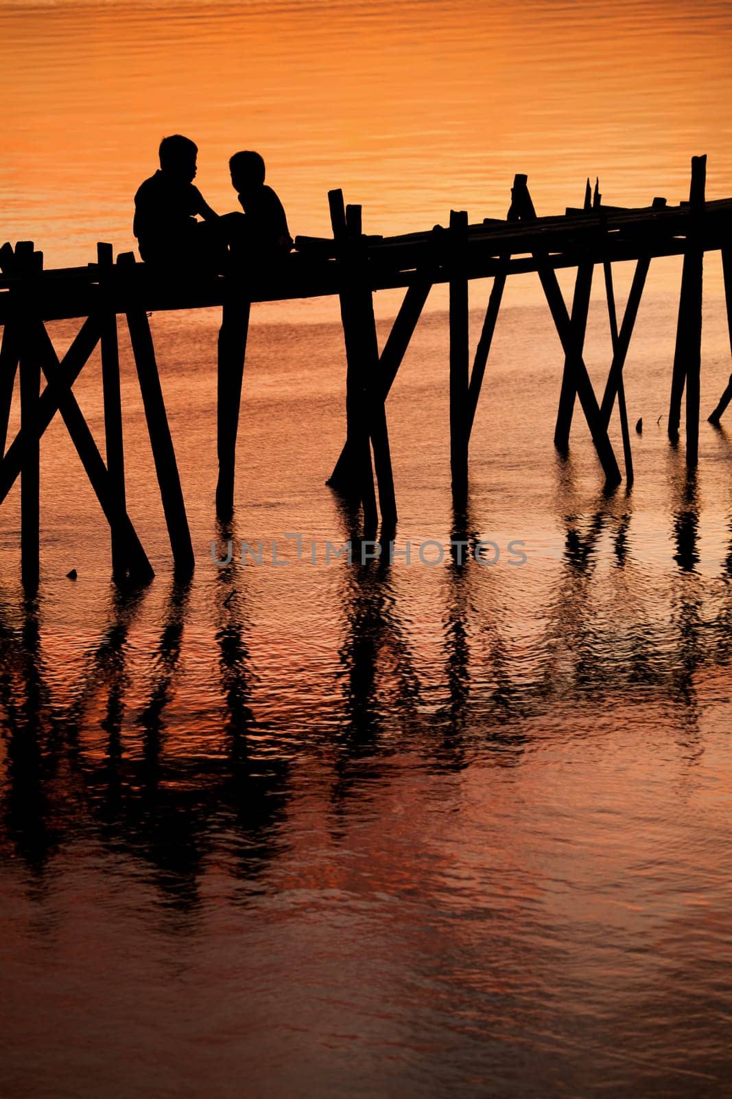 Silhouette of two children on an old fragile wooden bridge at sunset.