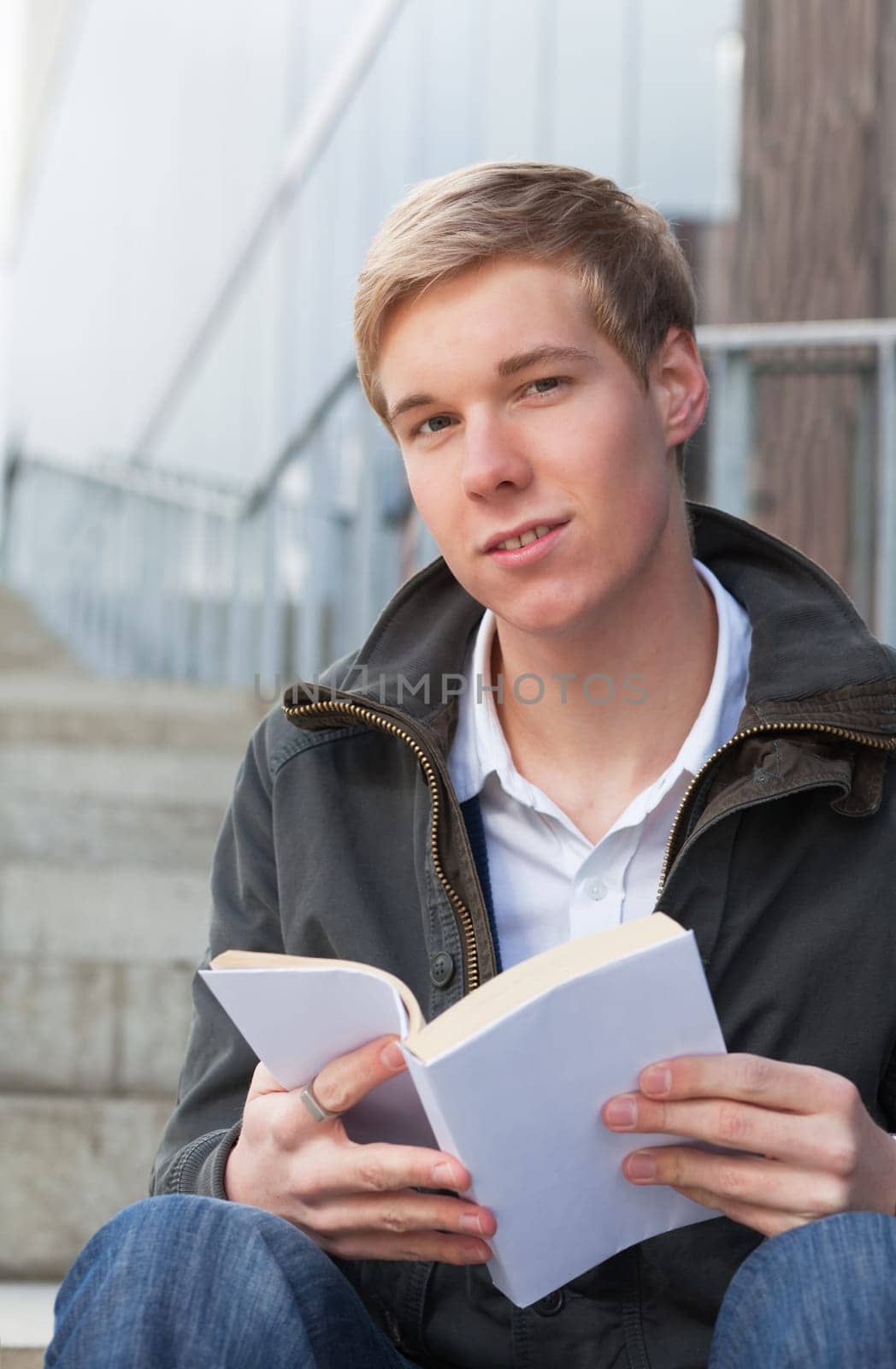 young blond handsome man sitting on stairs outdoors with book