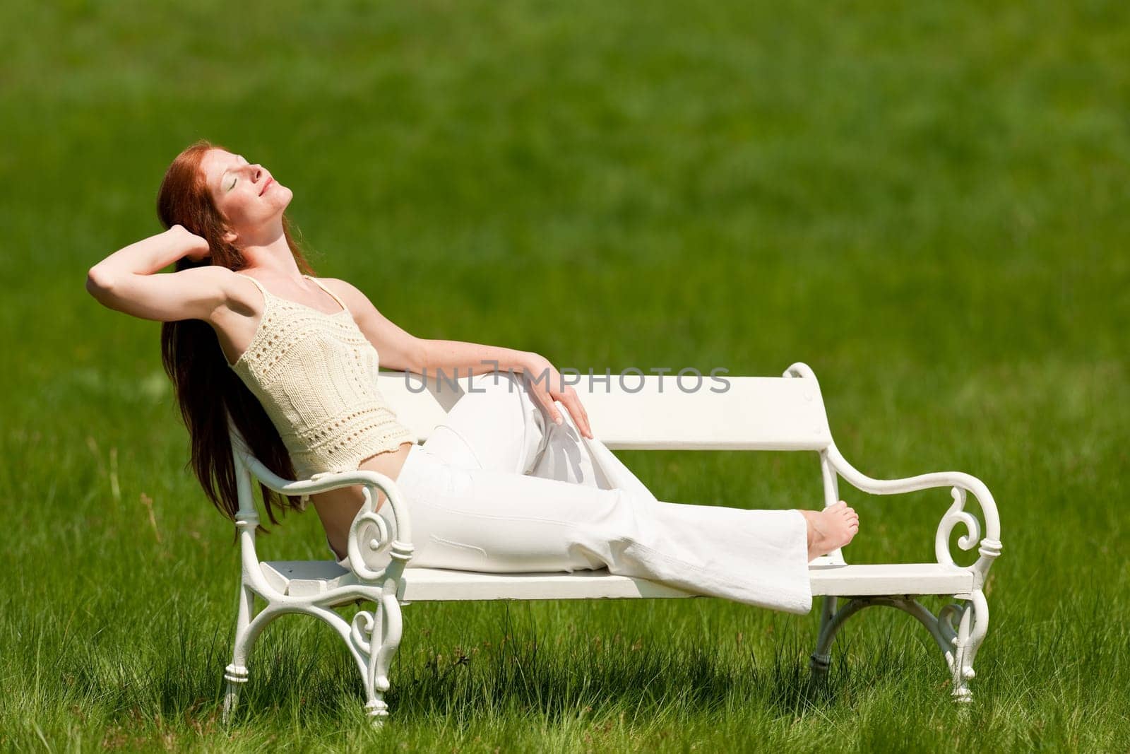 Red hair woman enjoying sun on white bench in a meadow shallow DOF