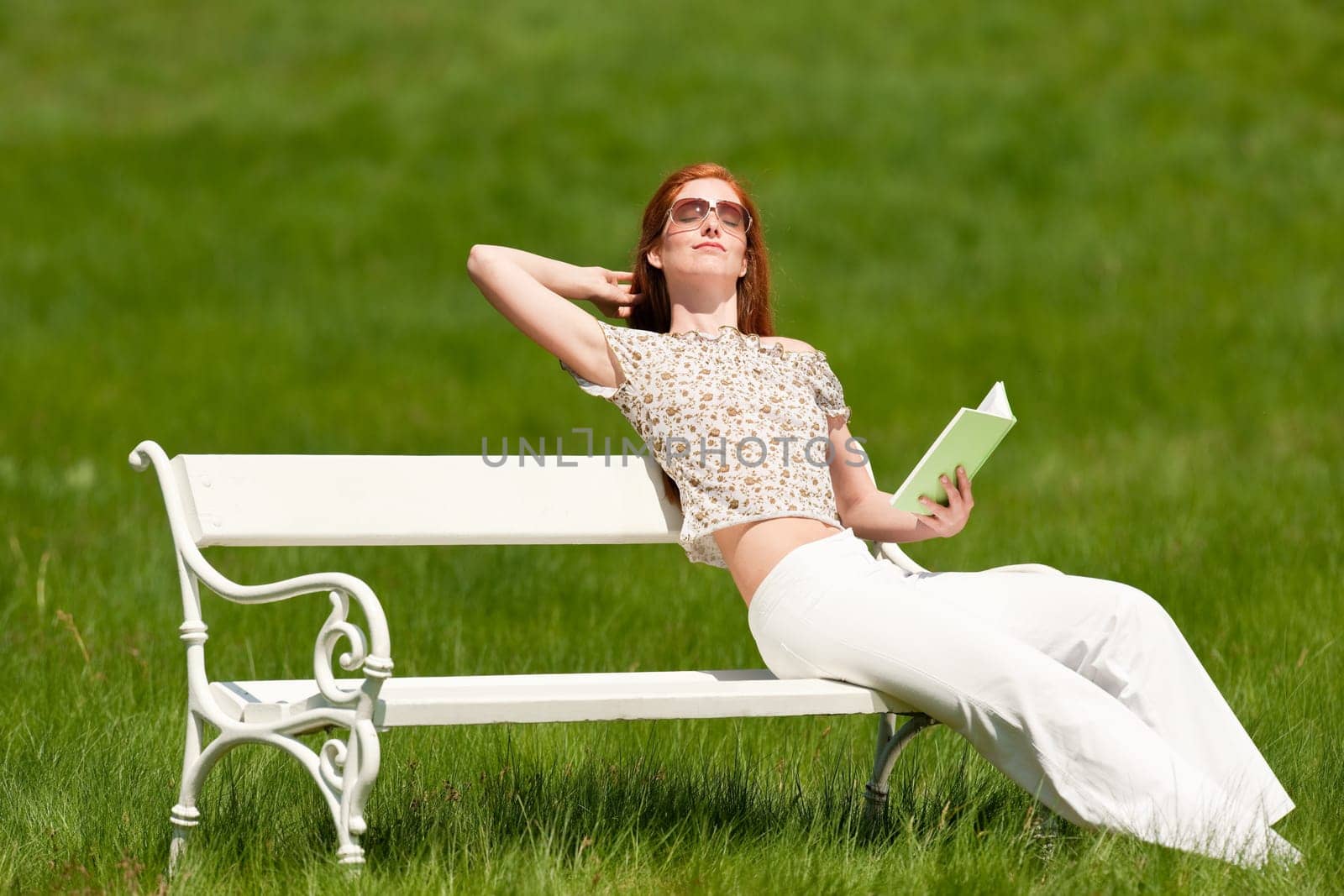 Red hair woman sitting with book on white bench in a meadow shallow DOF